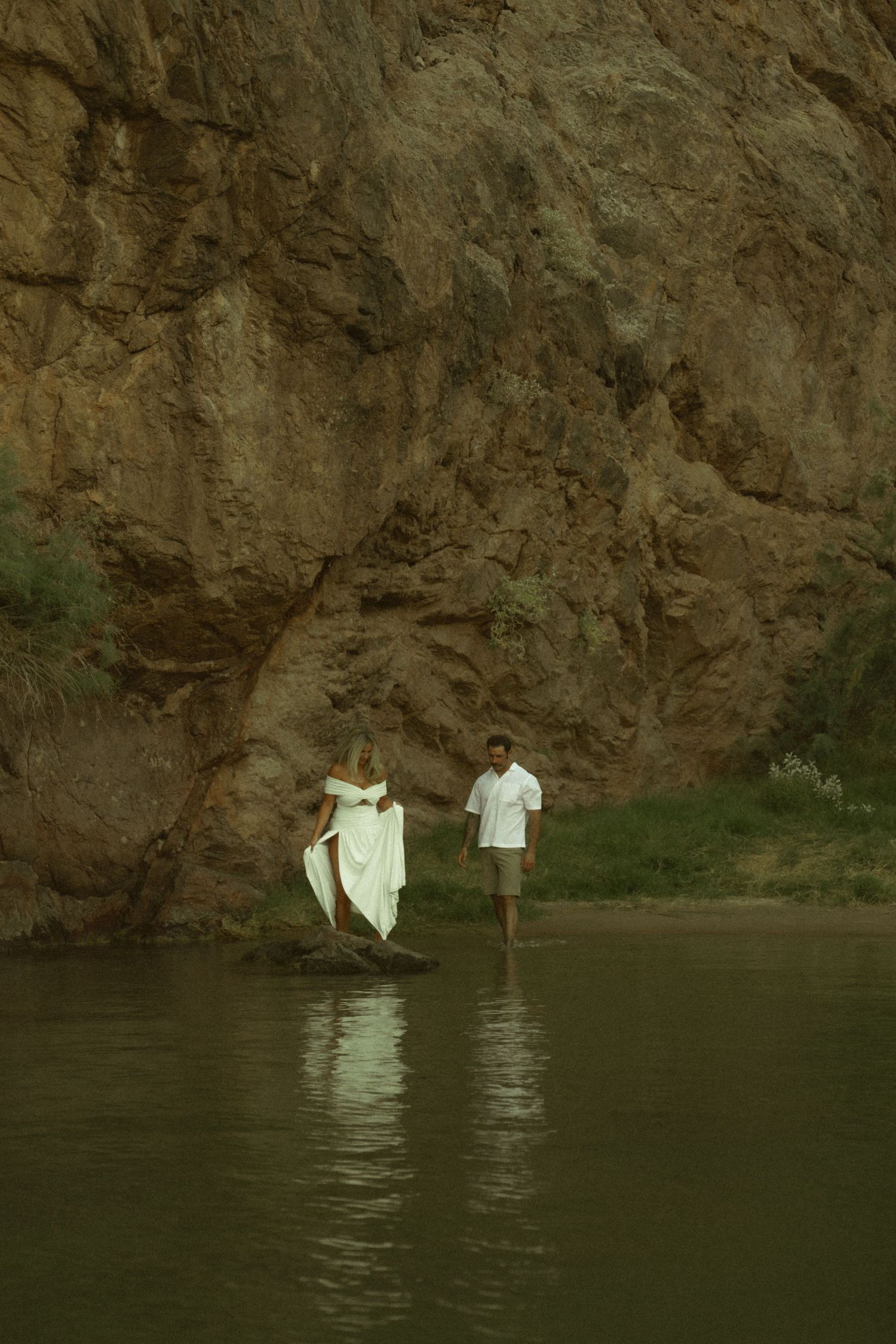 A couple in white attire stands by a rocky canyon river, the woman in a flowing gown and man wading in shallow water.