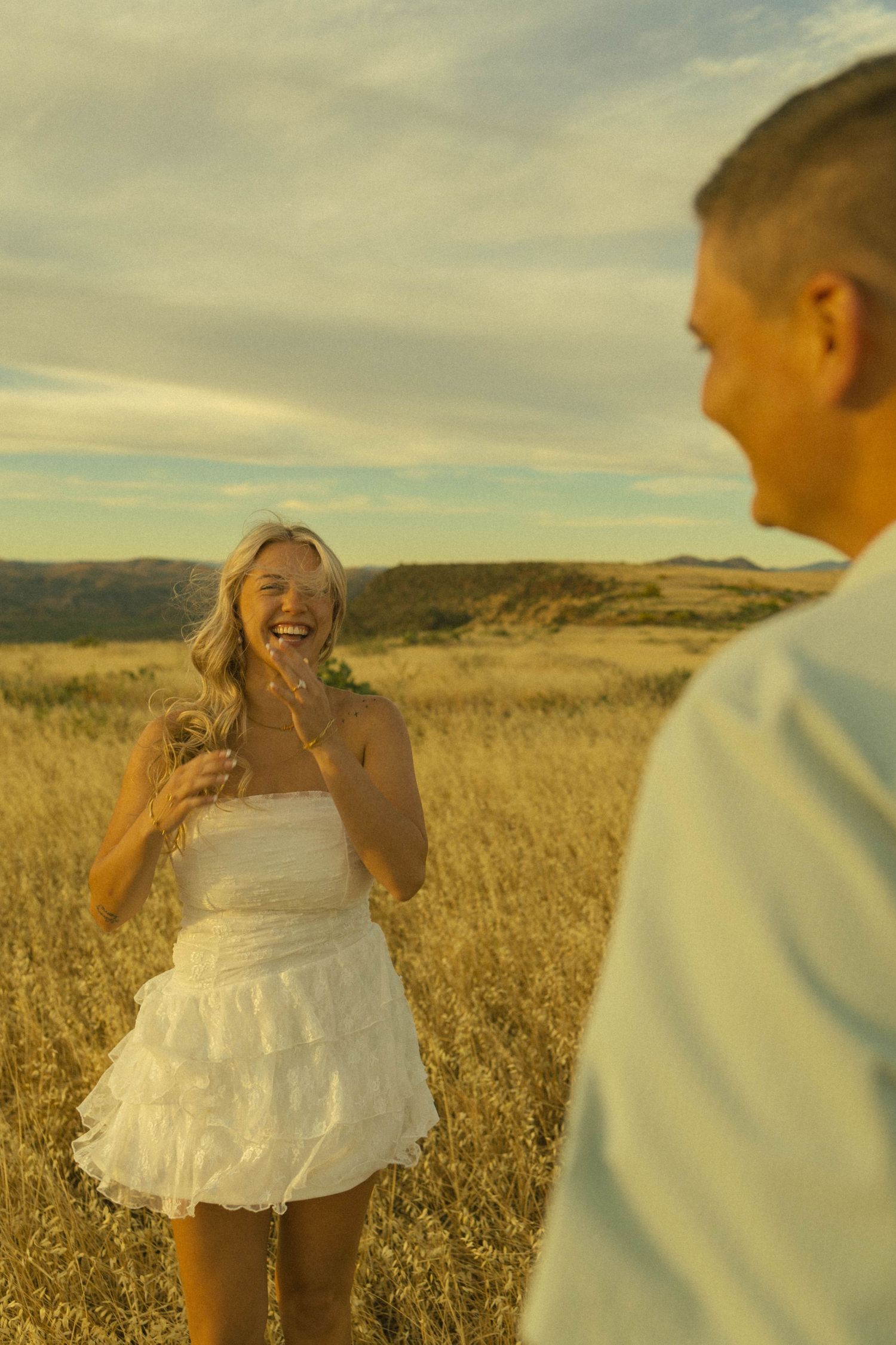 Laughing blonde woman in white dress stands in golden field at sunset with man in foreground.