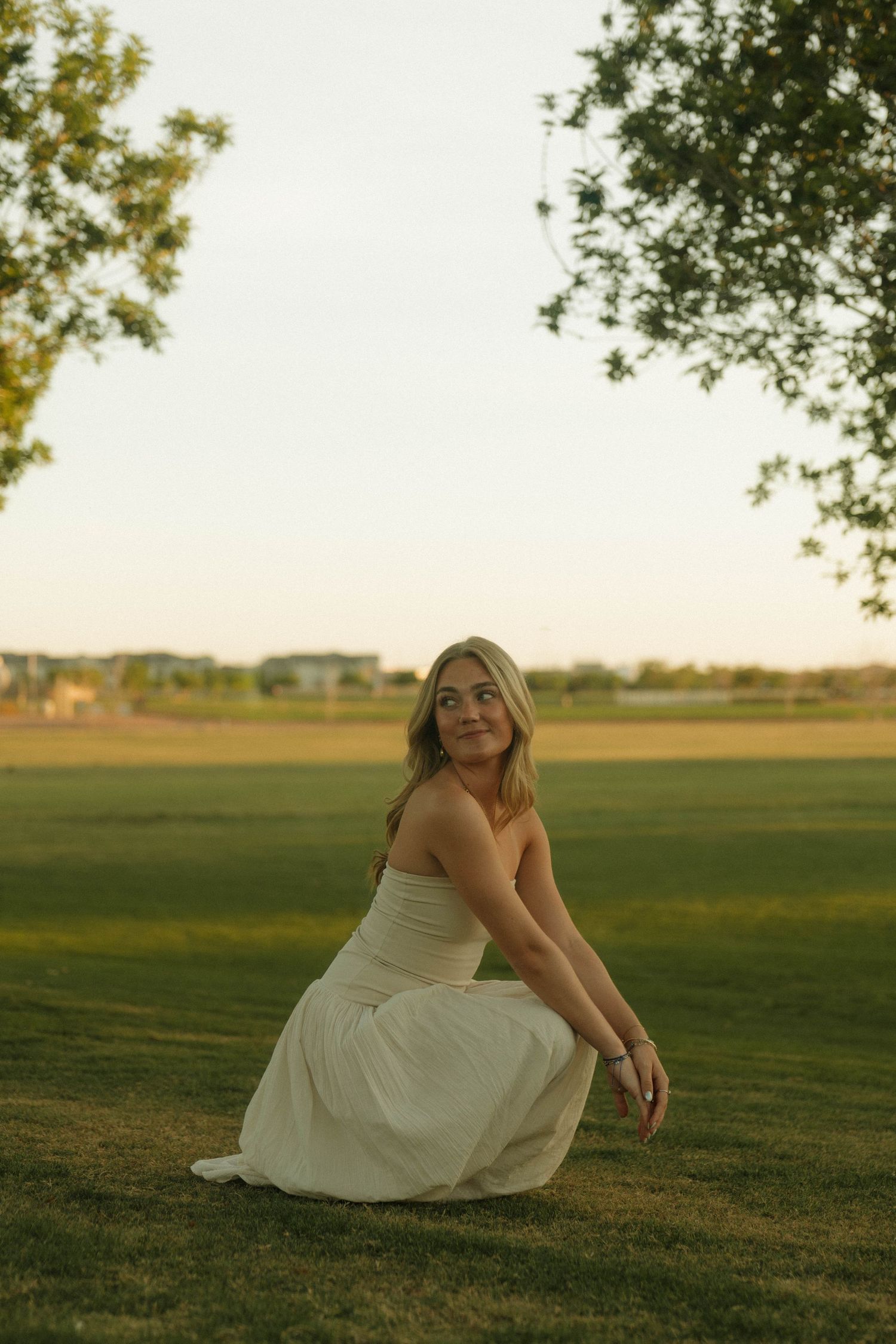 A young blonde woman in a white strapless dress crouches gracefully on green grass at golden hour.