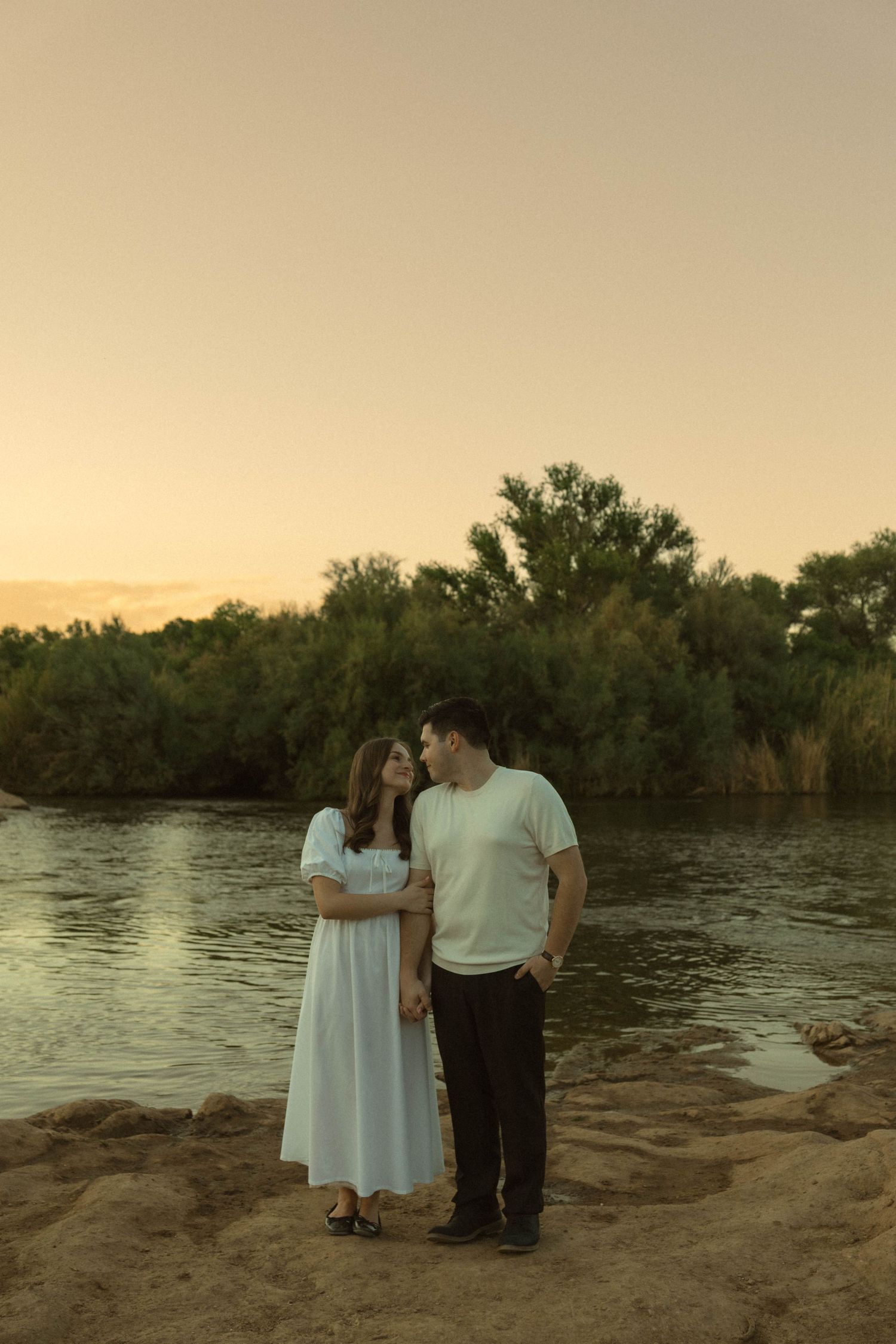Romantic couple standing together by river at sunset in warm golden light.