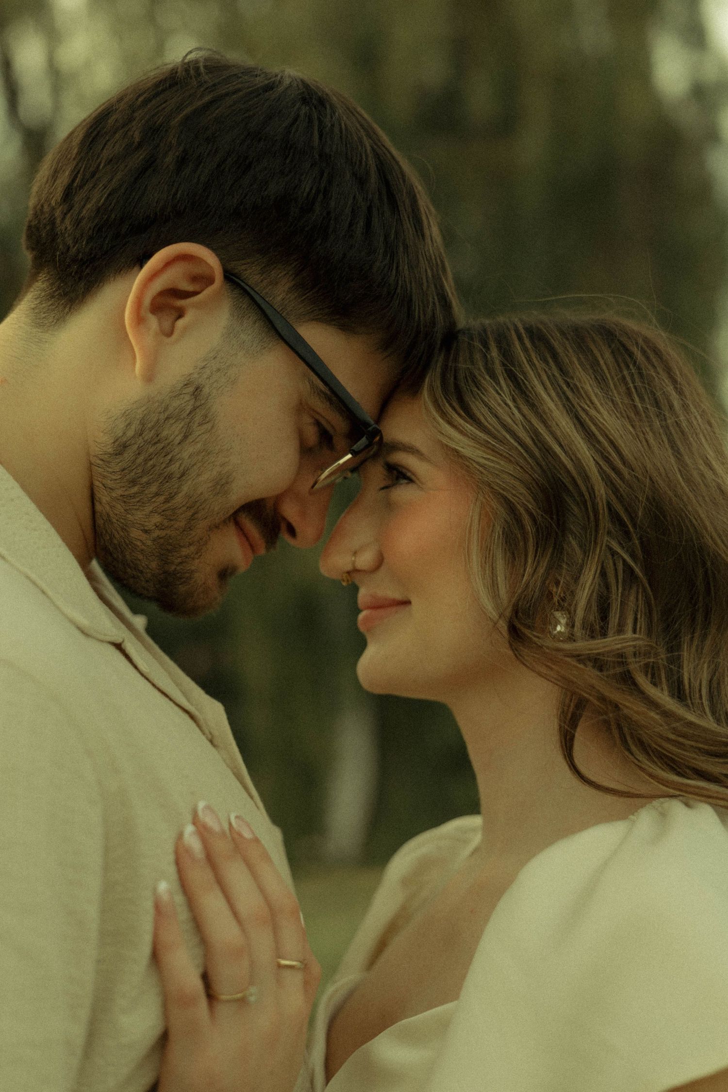 Romantic couple touching foreheads and smiling closely, man wearing glasses outdoors.
