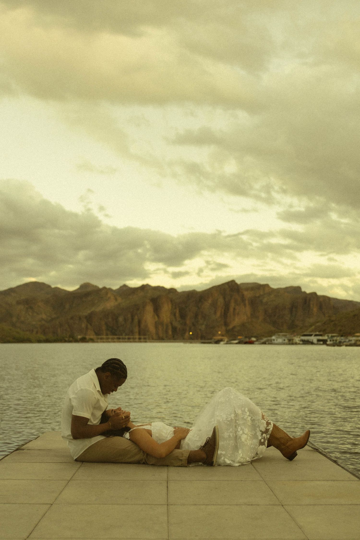 Romantic couple sharing an intimate moment on a waterfront dock at sunset with dramatic mountain backdrop.