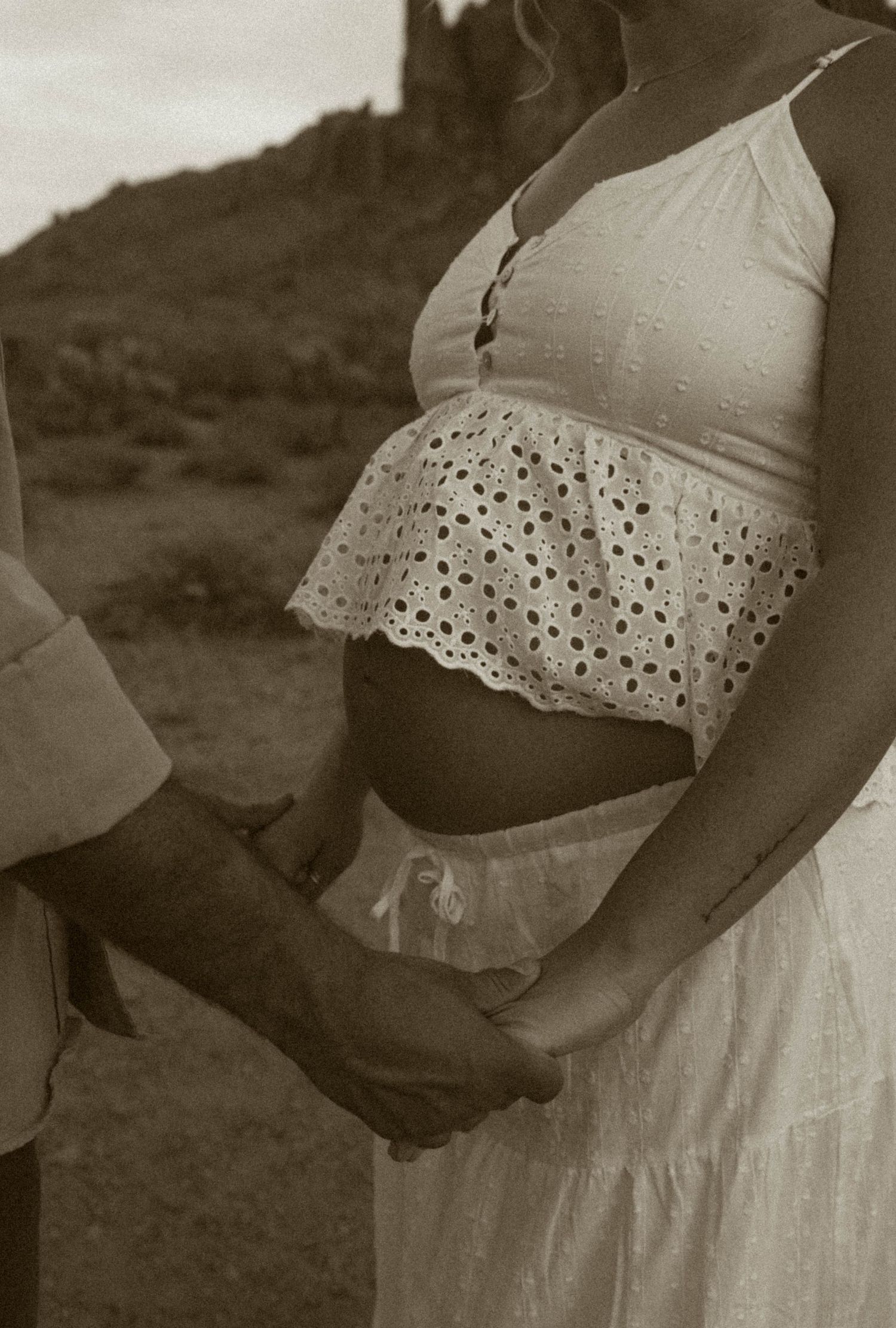 Close-up of pregnant woman holding hands with partner in desert setting, sepia tone.