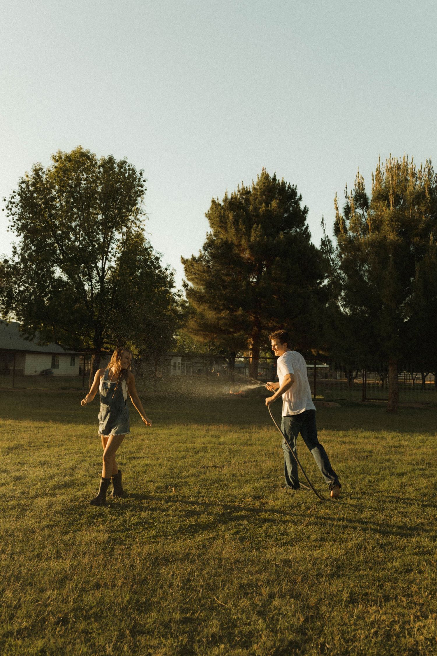 Playful couple chasing each other with a garden hose on a green lawn at golden hour.