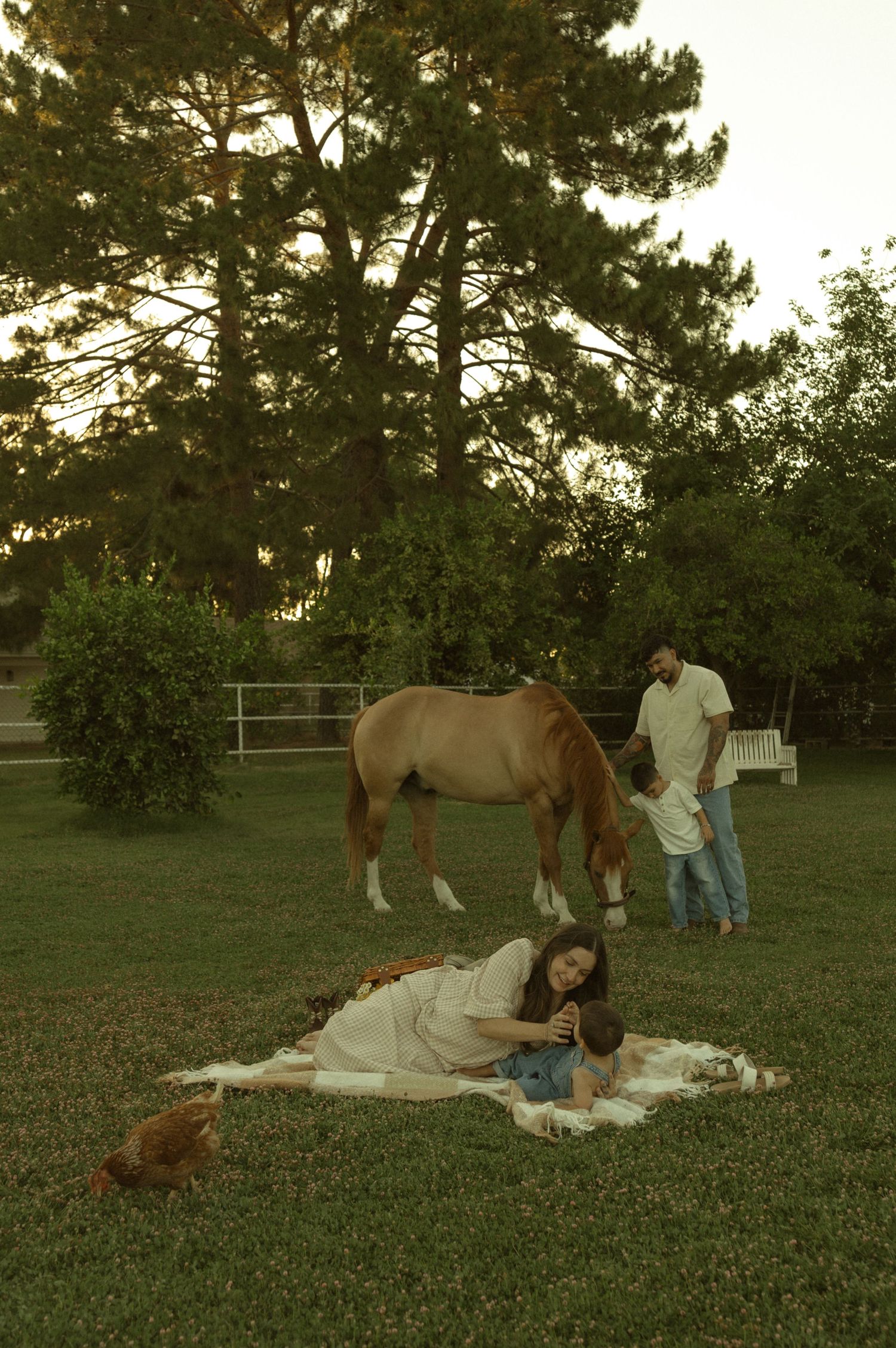 Family relaxing on a blanket in a lush garden with a chestnut horse standing nearby.