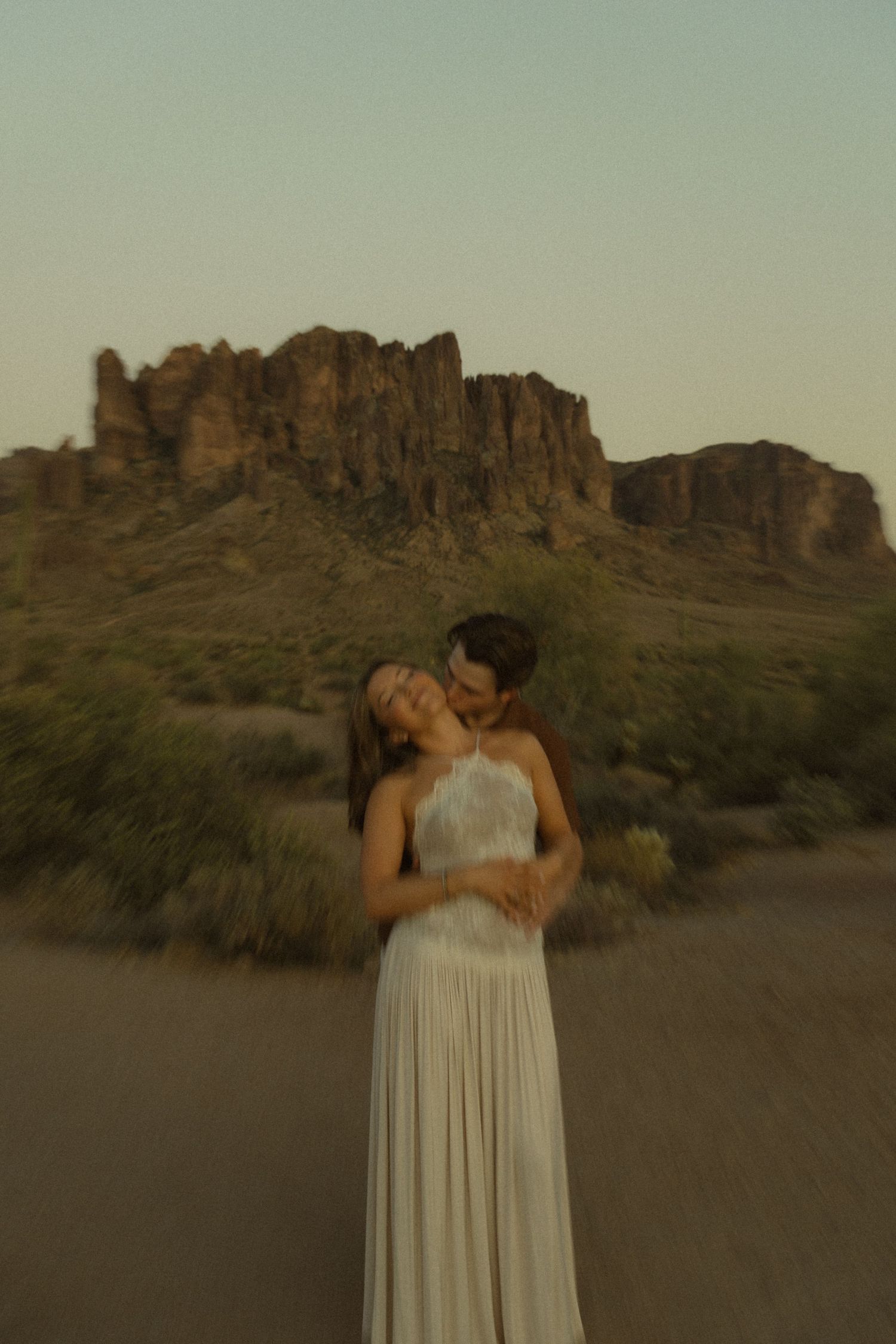 Man embracing woman in white halter dress from behind in soft-focus desert photo with mesa backdrop.