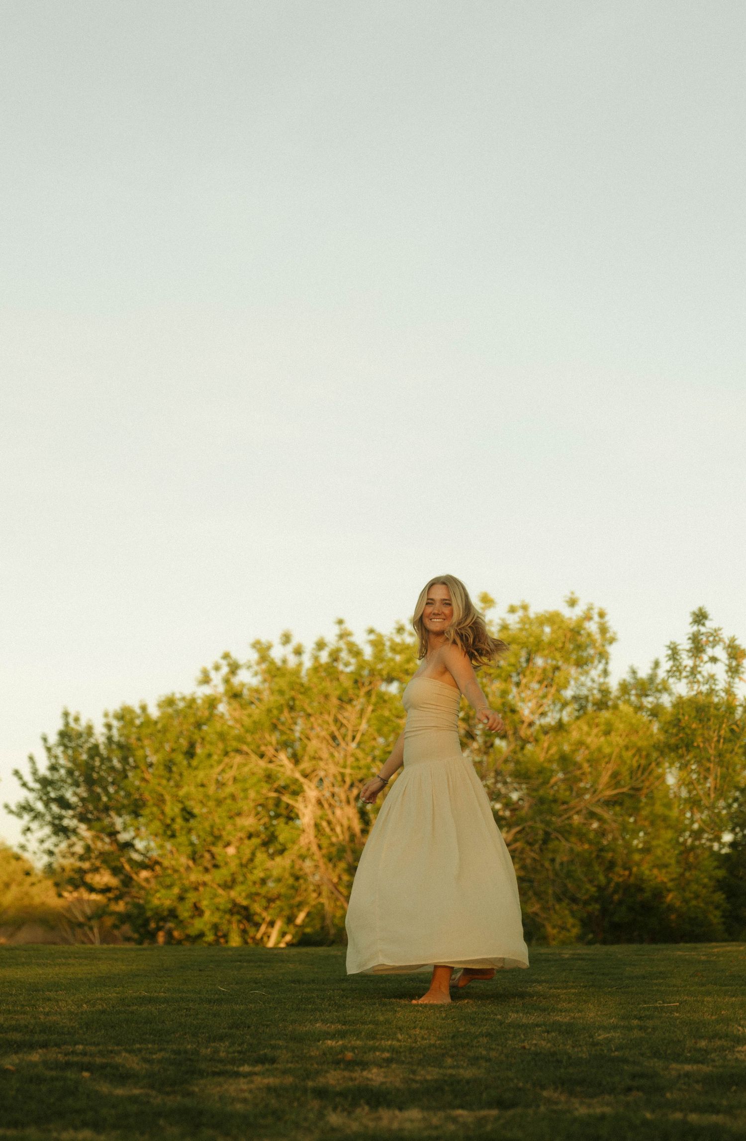 A smiling woman in a flowing cream maxi dress twirls on green grass at golden hour with trees in the background.