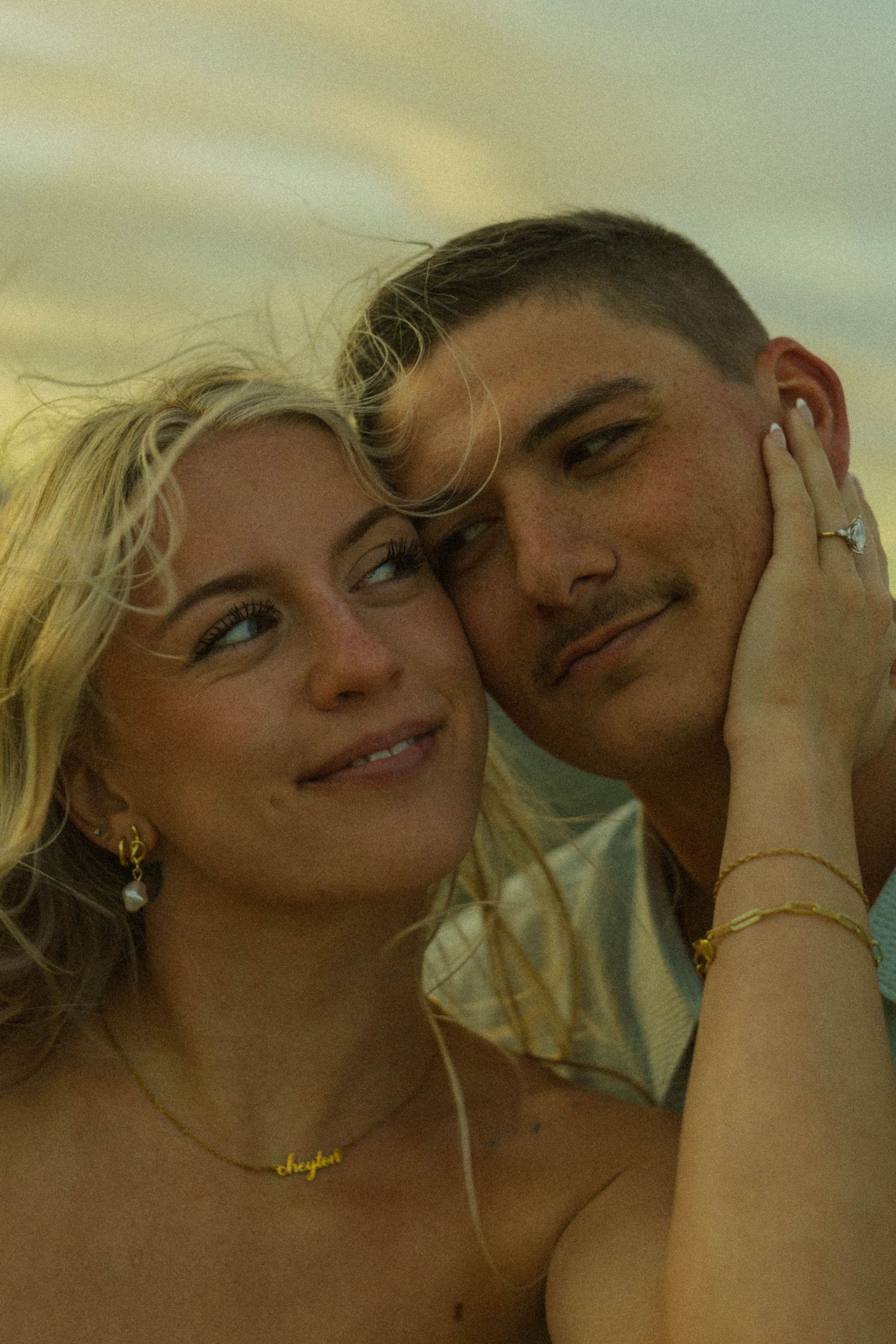 A young couple poses cheek to cheek outdoors at golden hour, woman with blonde hair wearing gold jewelry.