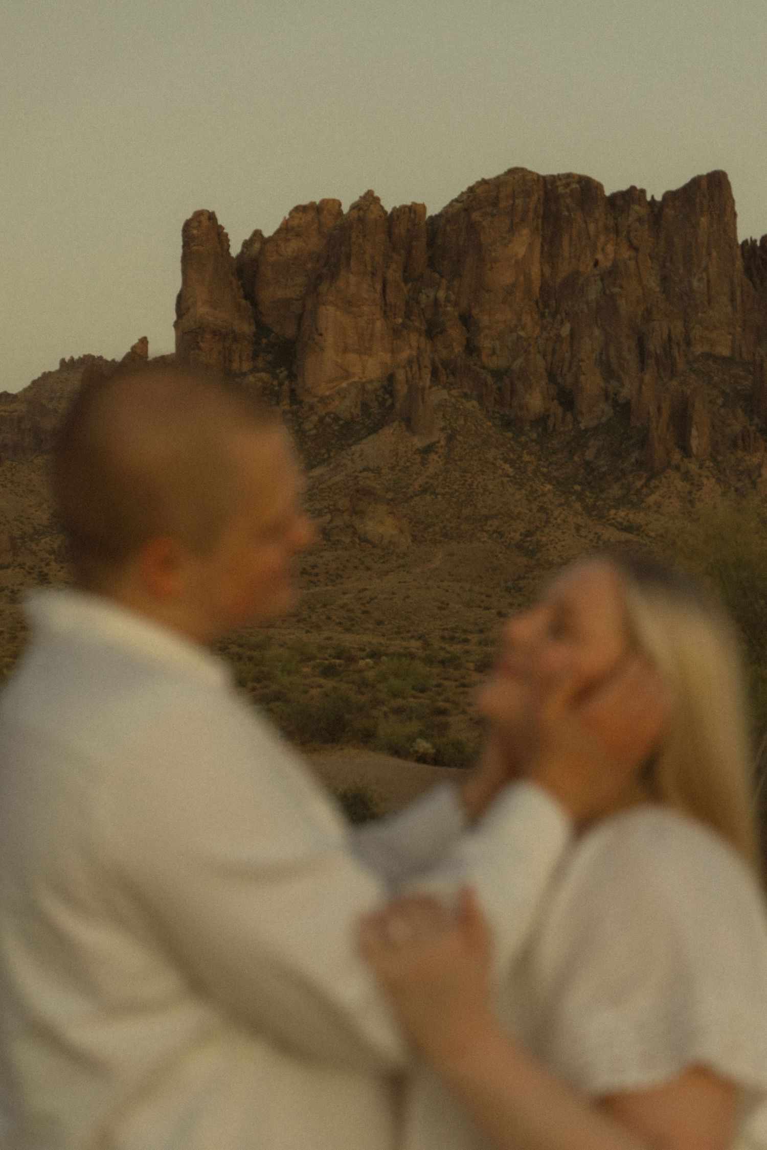 Couple in white embracing at sunset with Superstition Mountains rocky desert landscape in background.