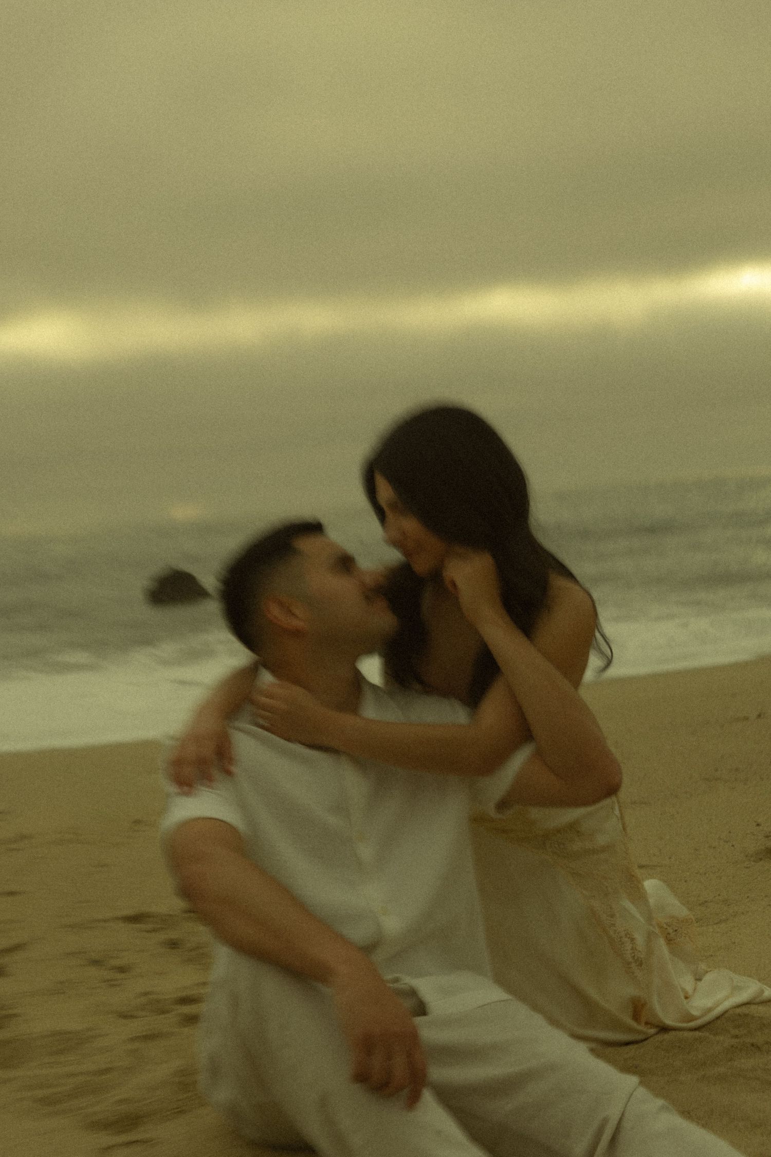Couple sharing a romantic moment on a sandy beach at dusk, sitting together with ocean waves in the background.