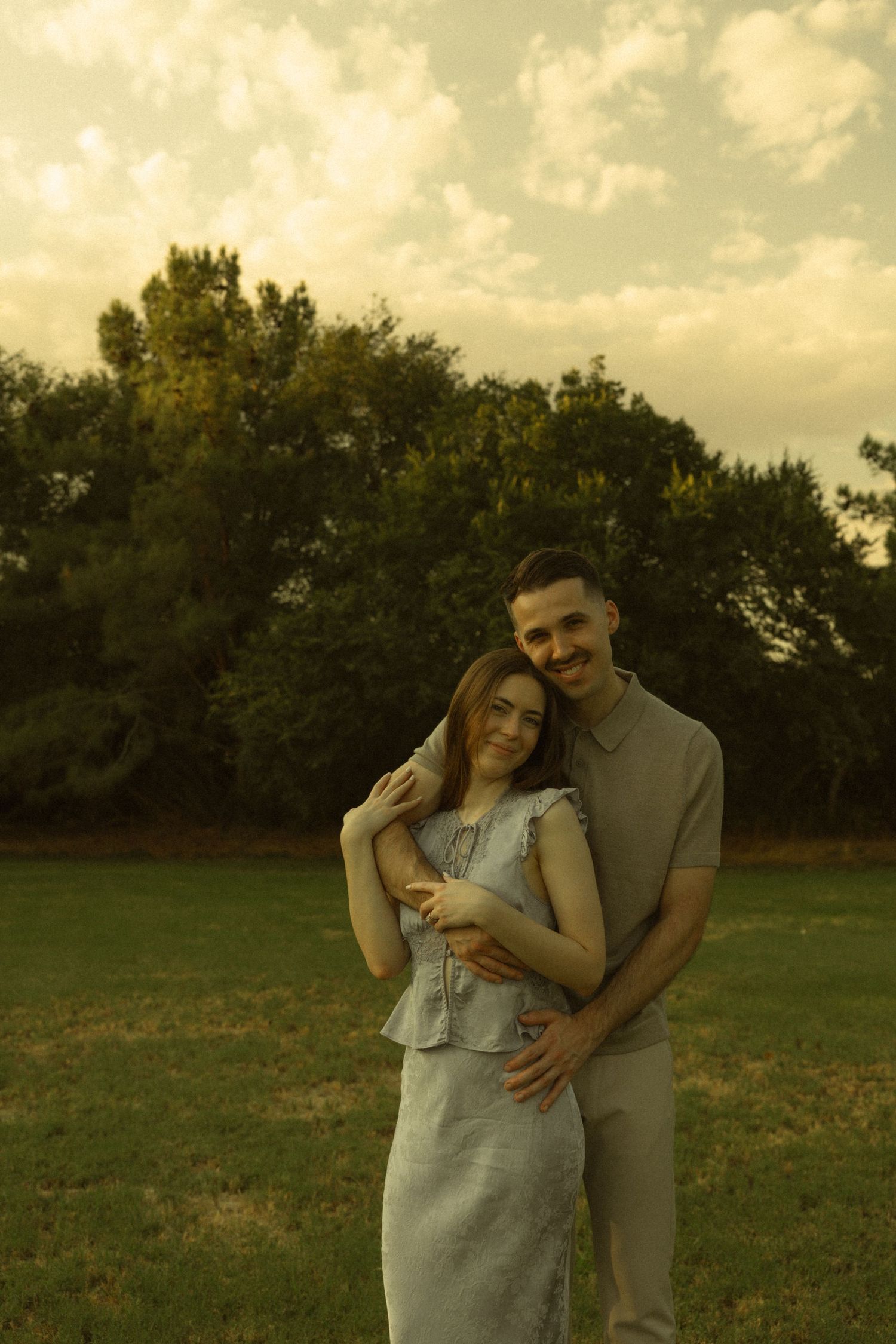 A couple embraces in a green park at golden hour, woman in gray dress leans back against man in beige polo.