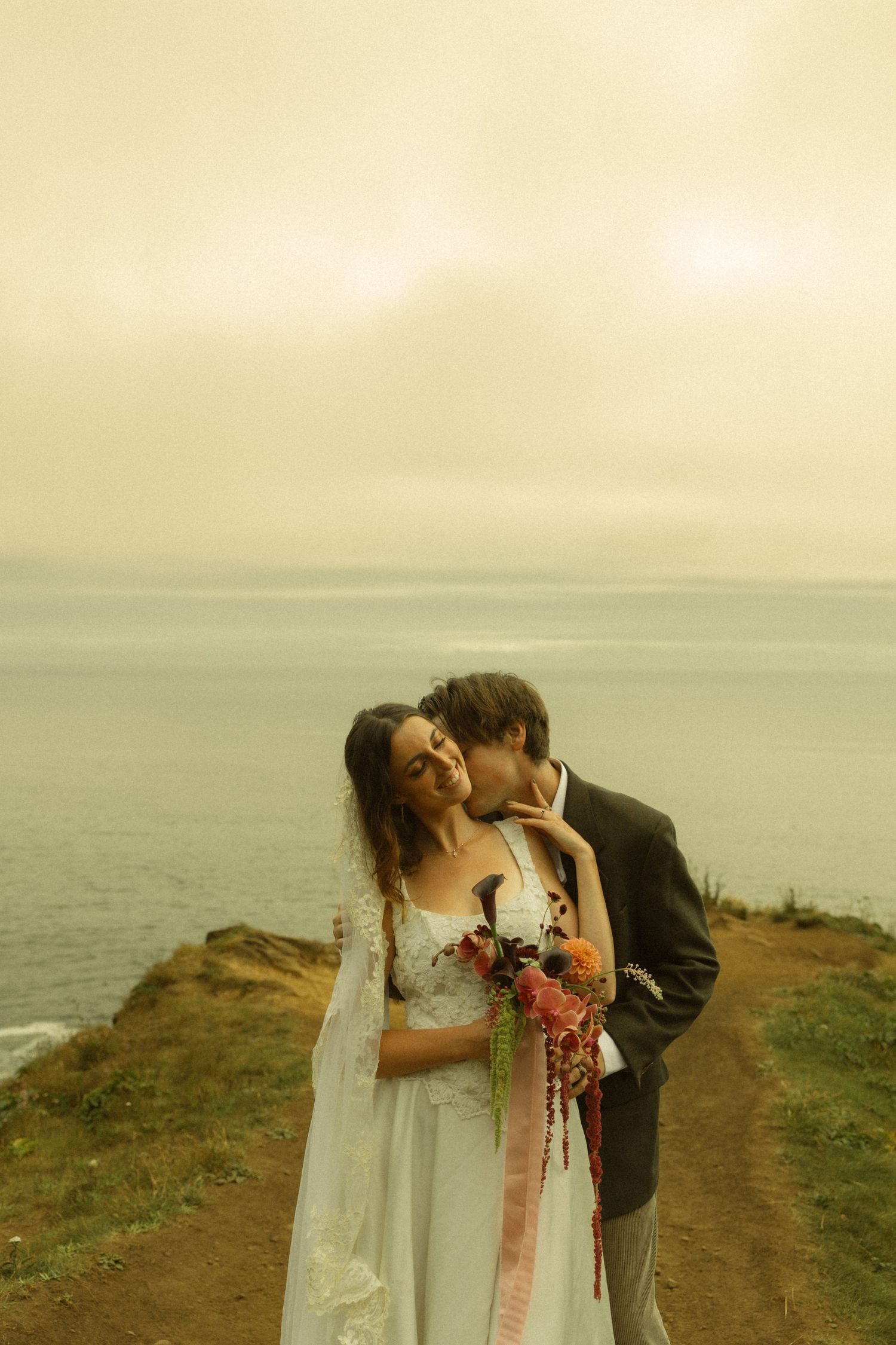 Bride and groom share a romantic kiss on a coastal cliff, with the ocean and golden sky in the background.