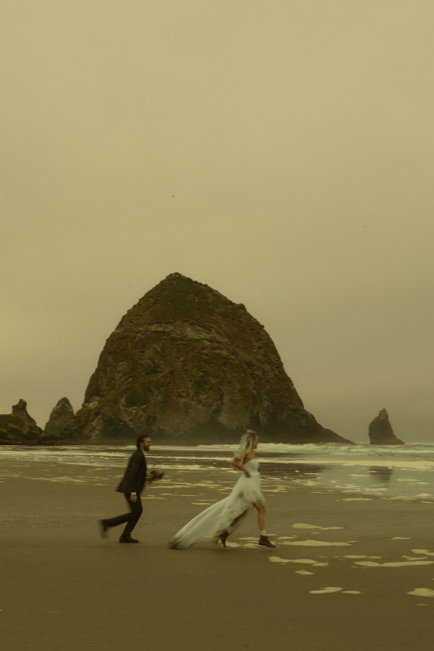 Newlyweds run playfully along the wet sand at Cannon Beach with Haystack Rock visible in the moody, overcast sky.