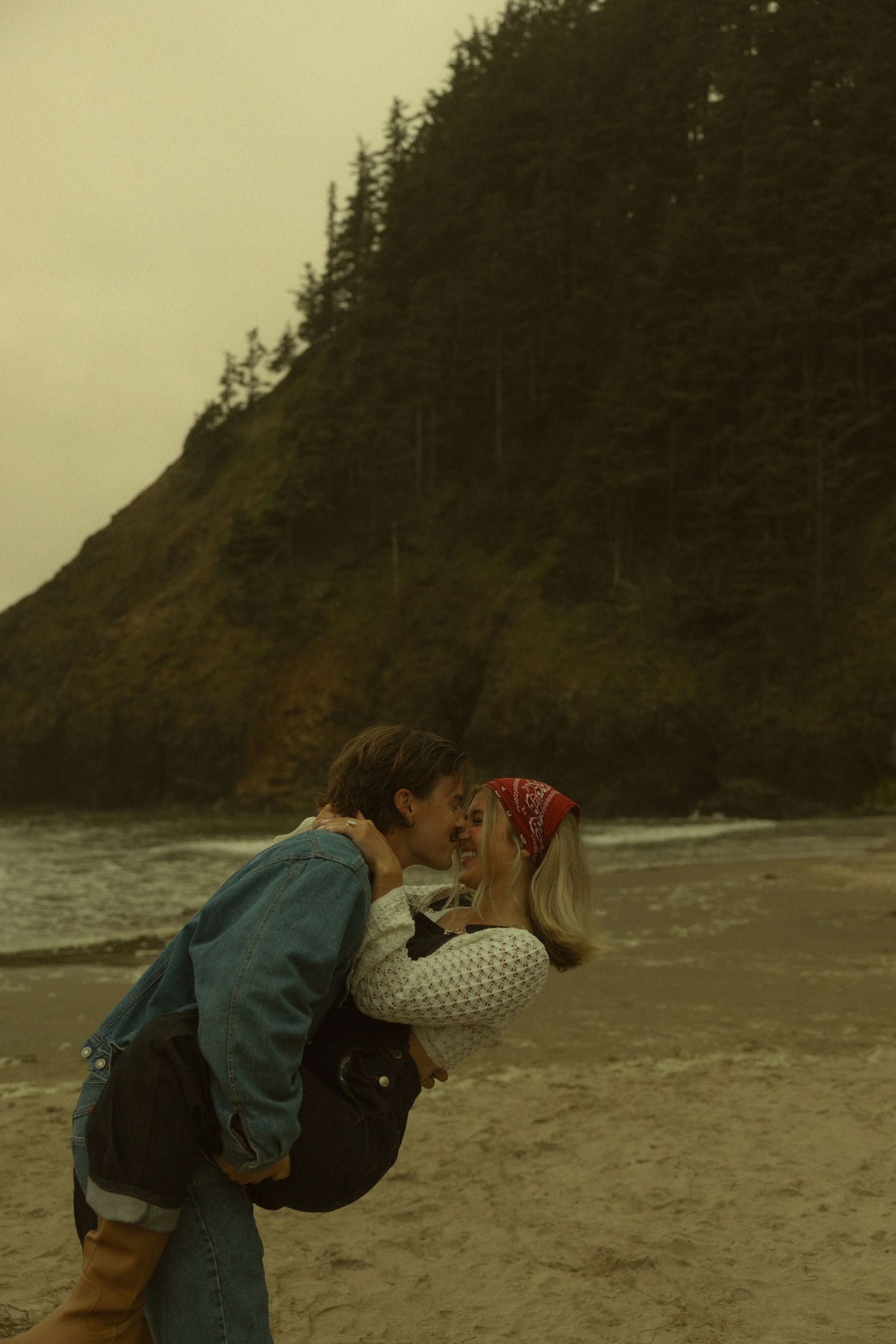 A man lifts a woman as they kiss on a sandy beach with a forested cliff and ocean waves behind them.