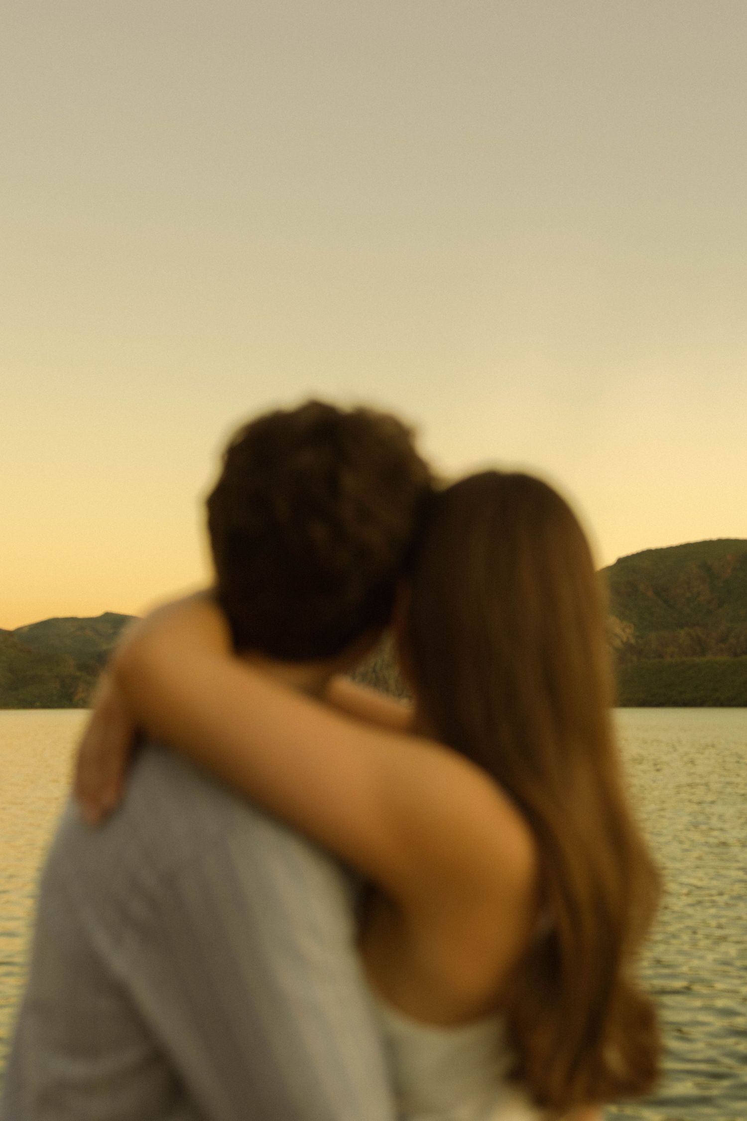 Two people share a romantic moment silhouetted against a serene lake and mountains at sunset.
