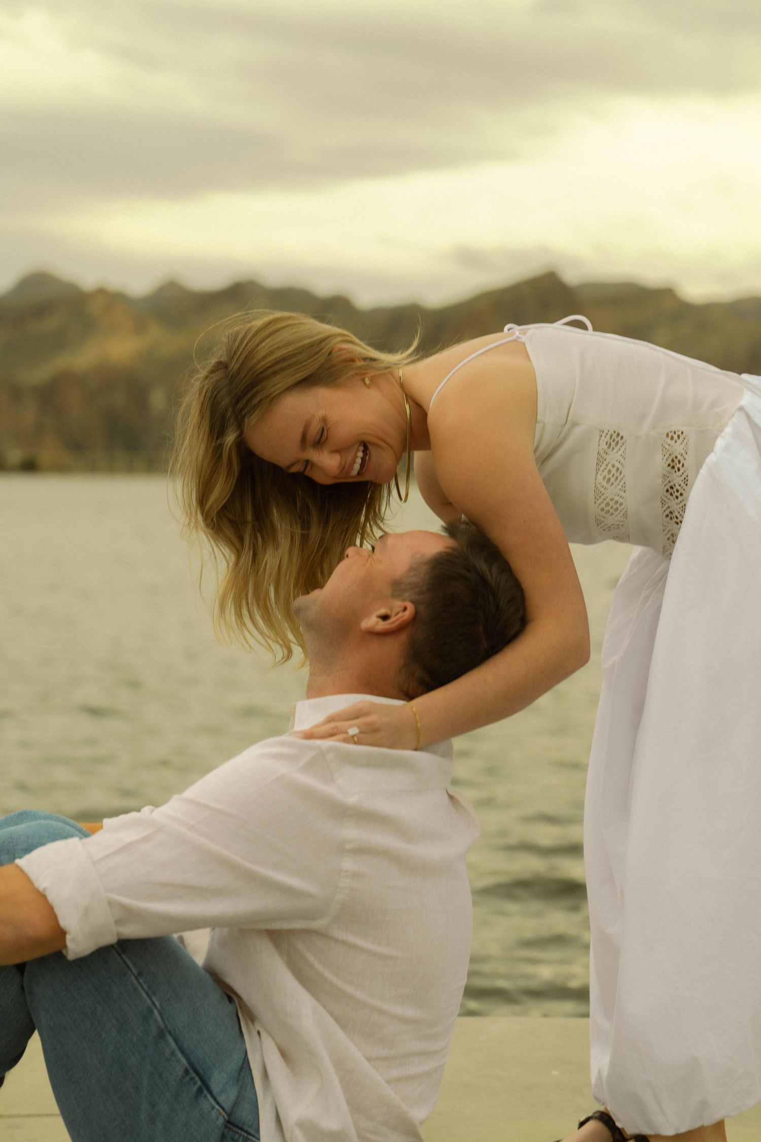 Playful couple in white outfits dancing and laughing by waterfront.