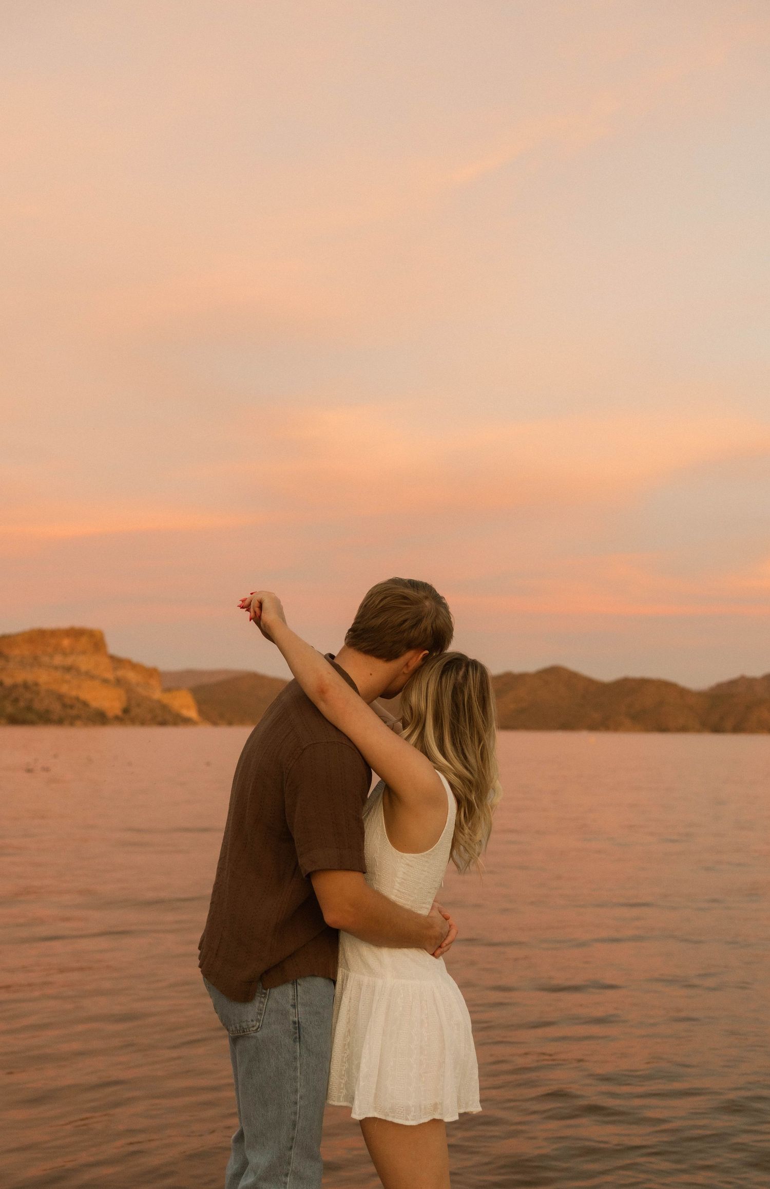 A couple embraces on a lakeside during a golden sunset with desert cliffs in the background.