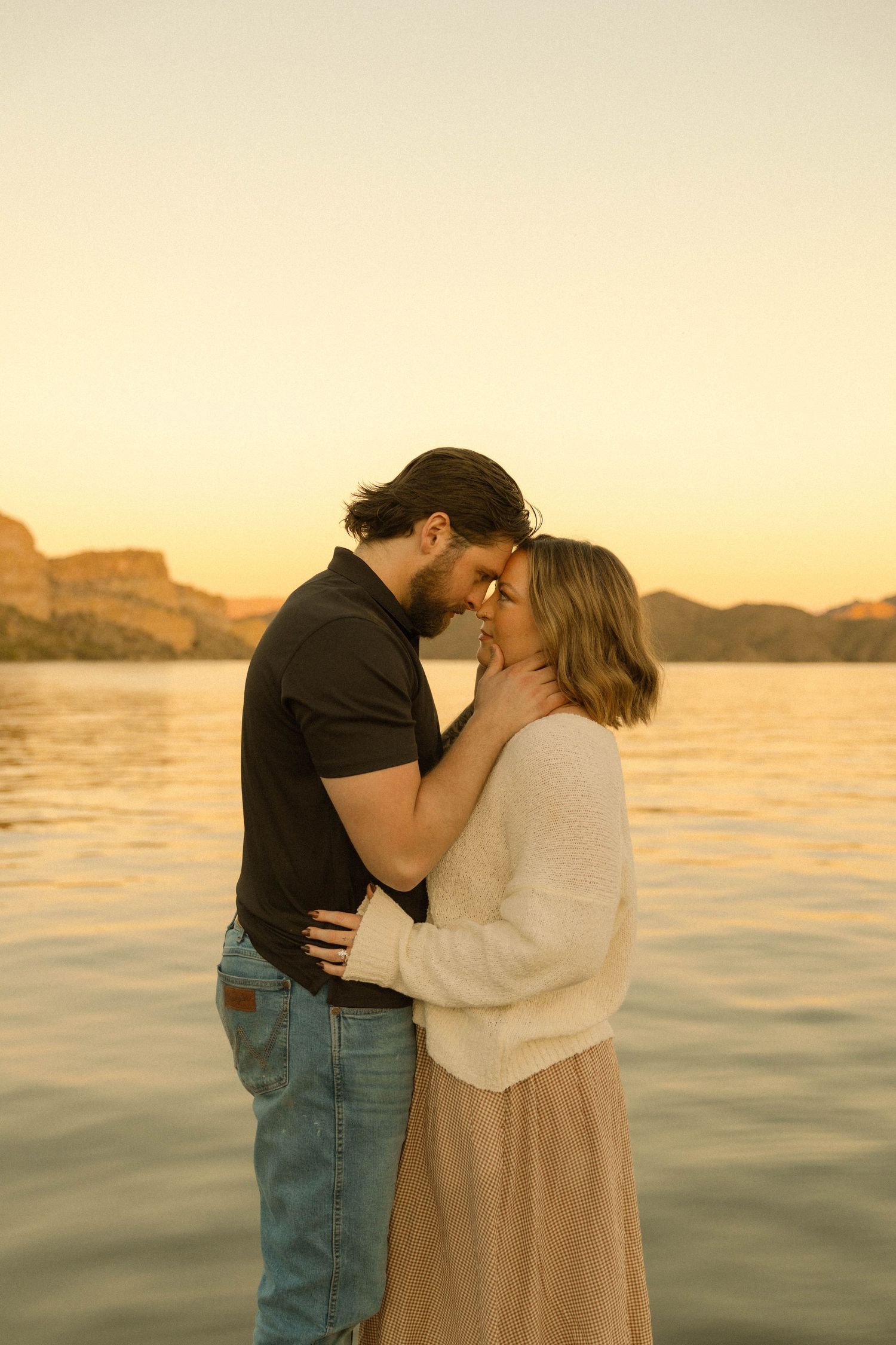A couple shares a romantic moment at sunset on a waterfront dock with mountains in the background.
