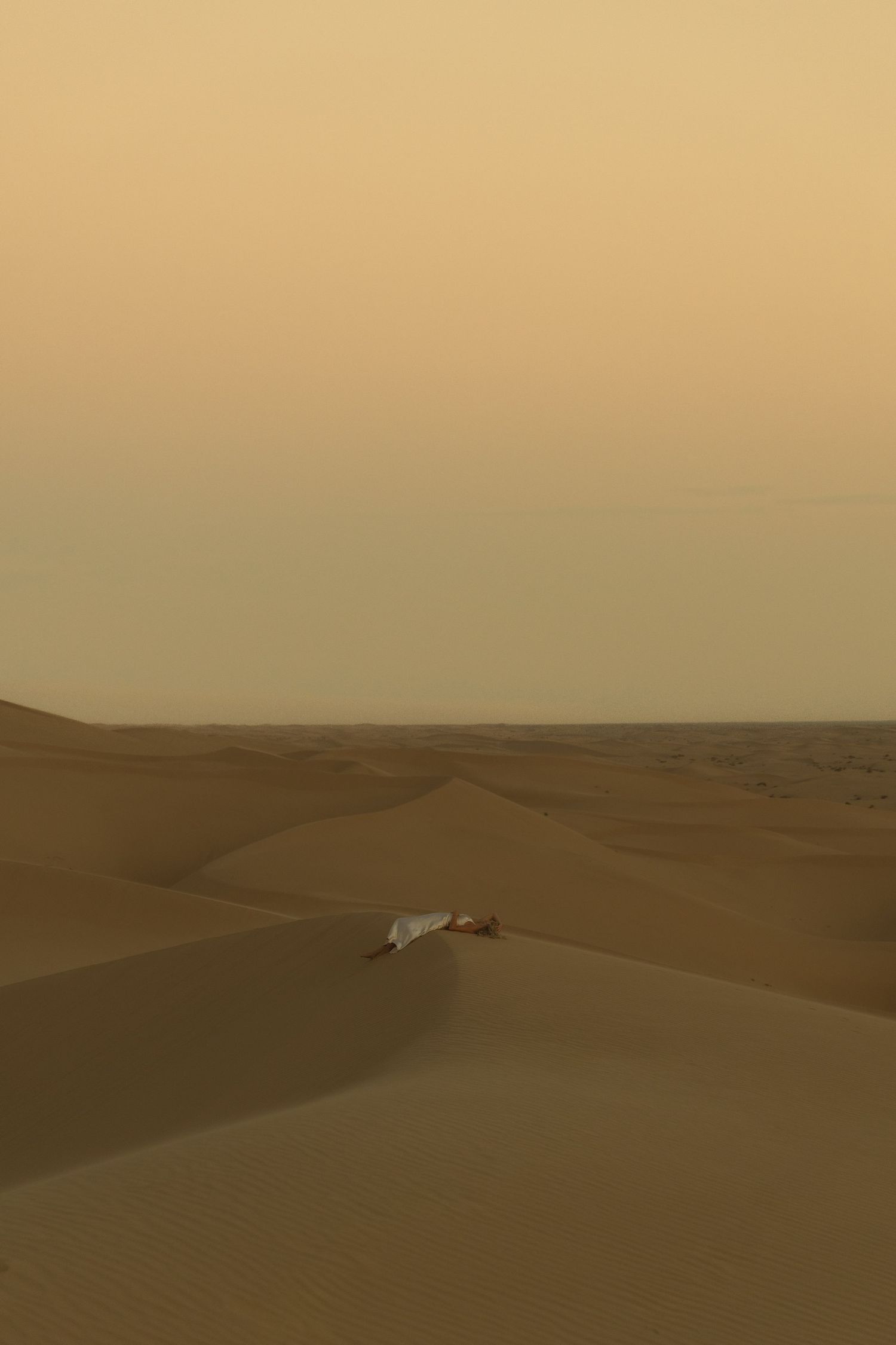 Lone figure resting on sweeping sand dunes in a vast desert landscape at dusk.