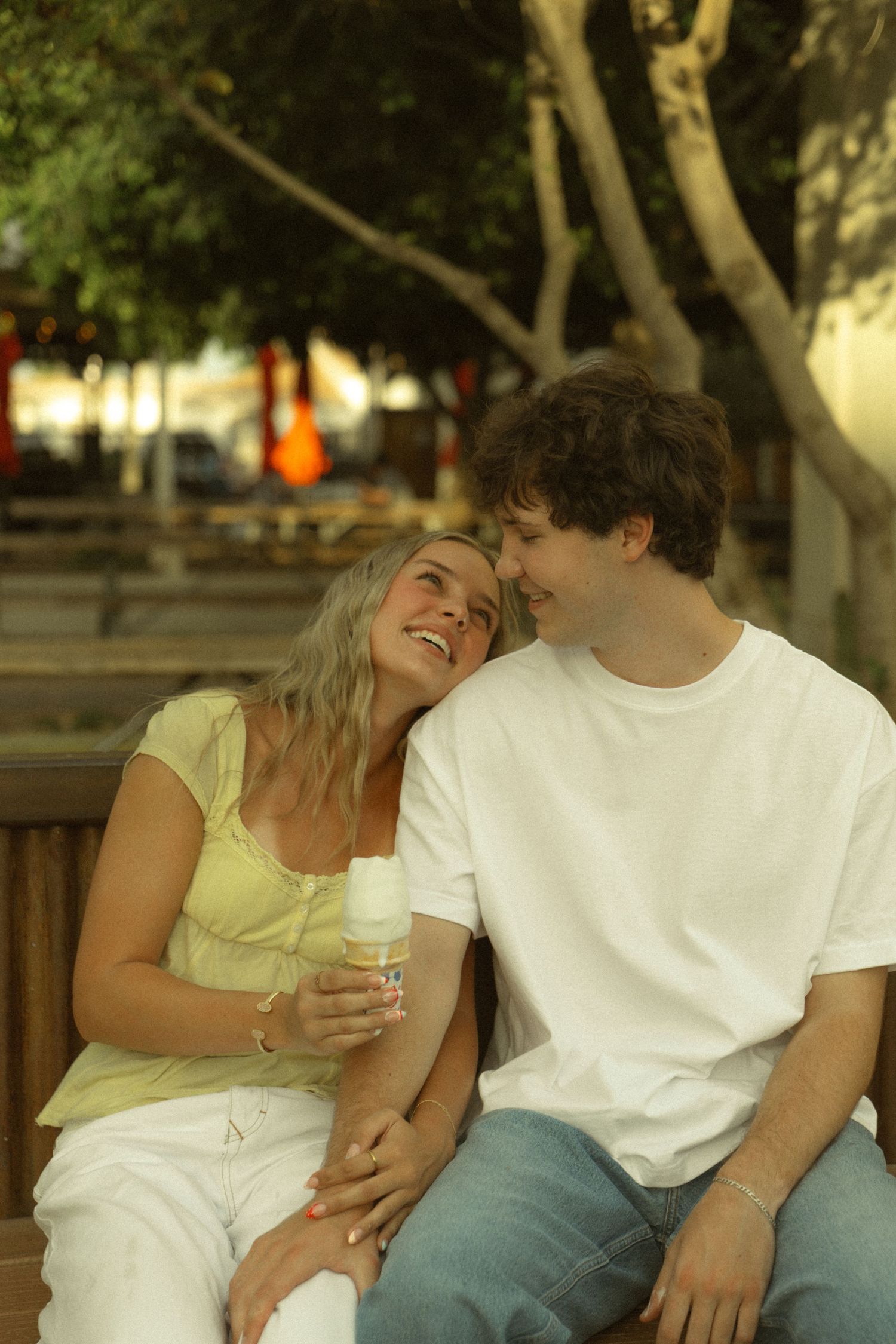 A happy young couple sitting on a bench outdoors, smiling at each other, woman holding a cold drink.
