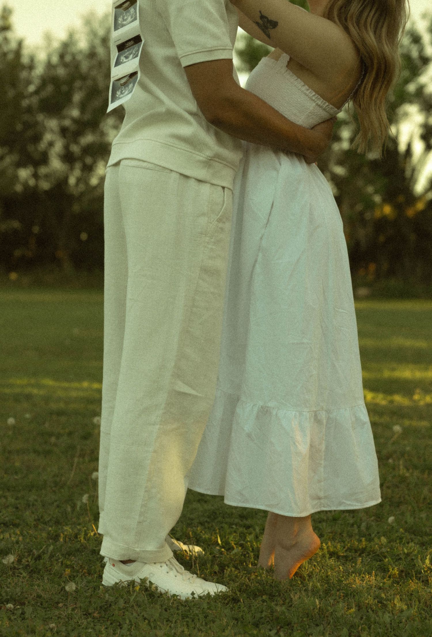A couple in white outfits embracing outdoors on green grass, holding ultrasound photos, suggesting a pregnancy announcement.