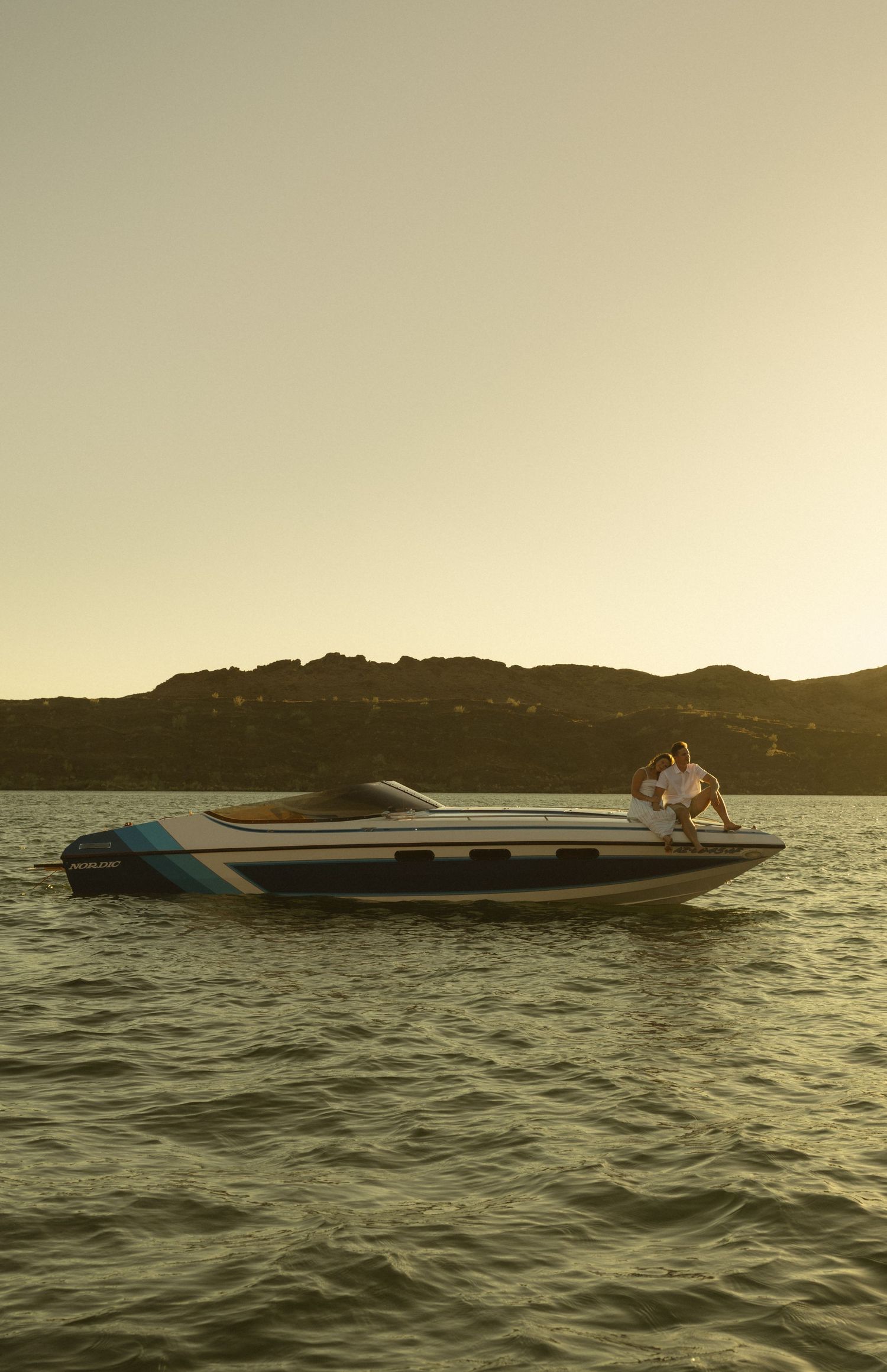 Two people relax on a sleek speedboat on calm waters at sunset with hills in the background.