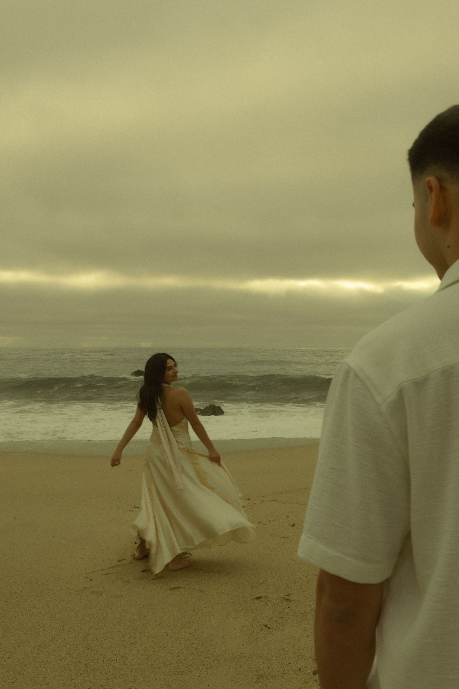 A woman in a flowing white backless dress walks on a misty beach while a man in white watches her.