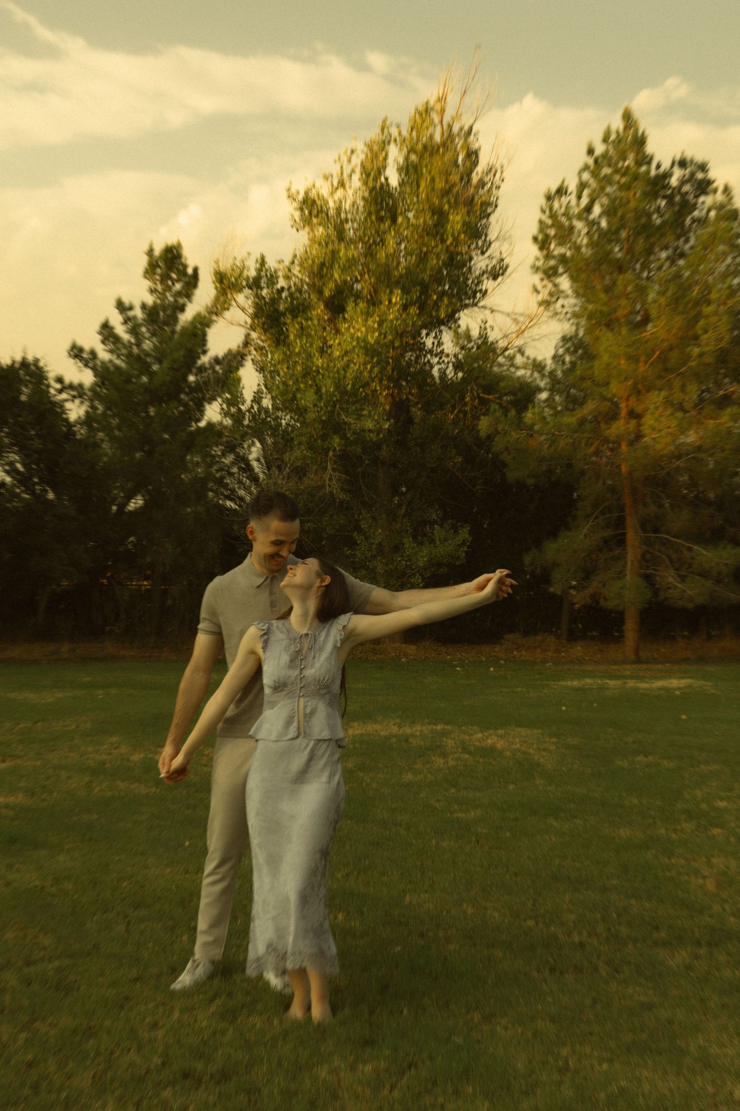 Couple dancing together on green grass in a park with lush trees and golden sky at sunset.
