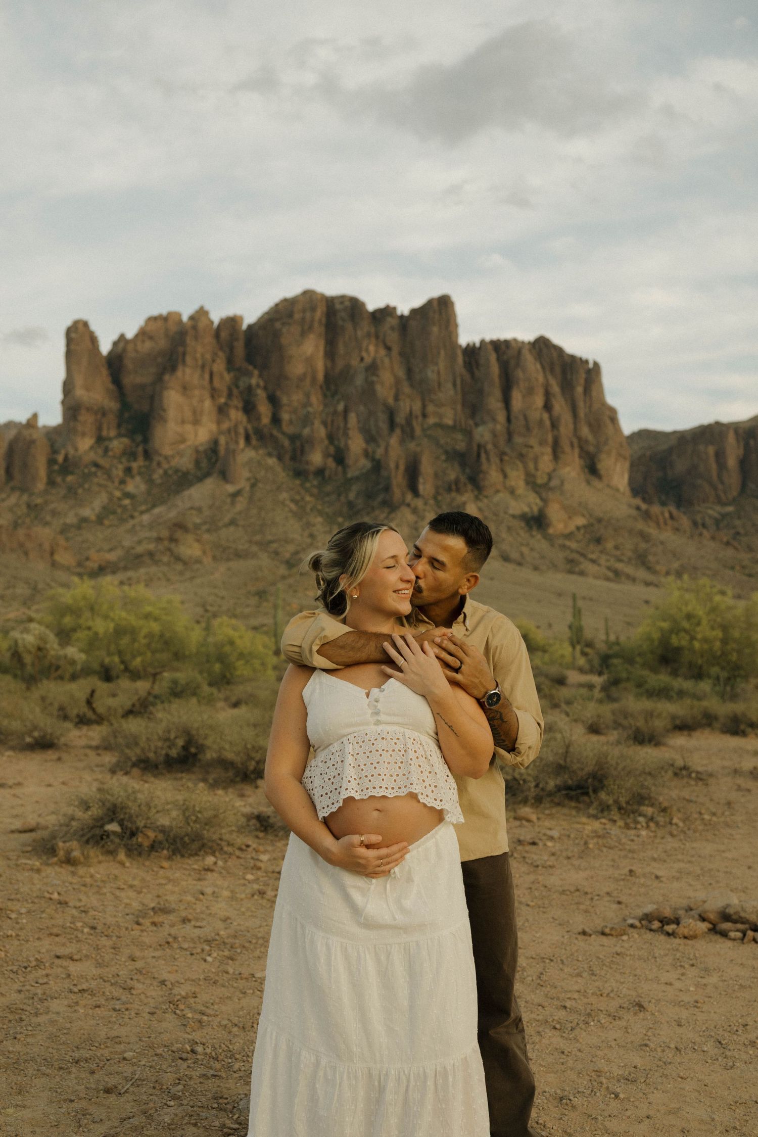 Man embraces pregnant woman in white dress with rocky desert mountains behind.