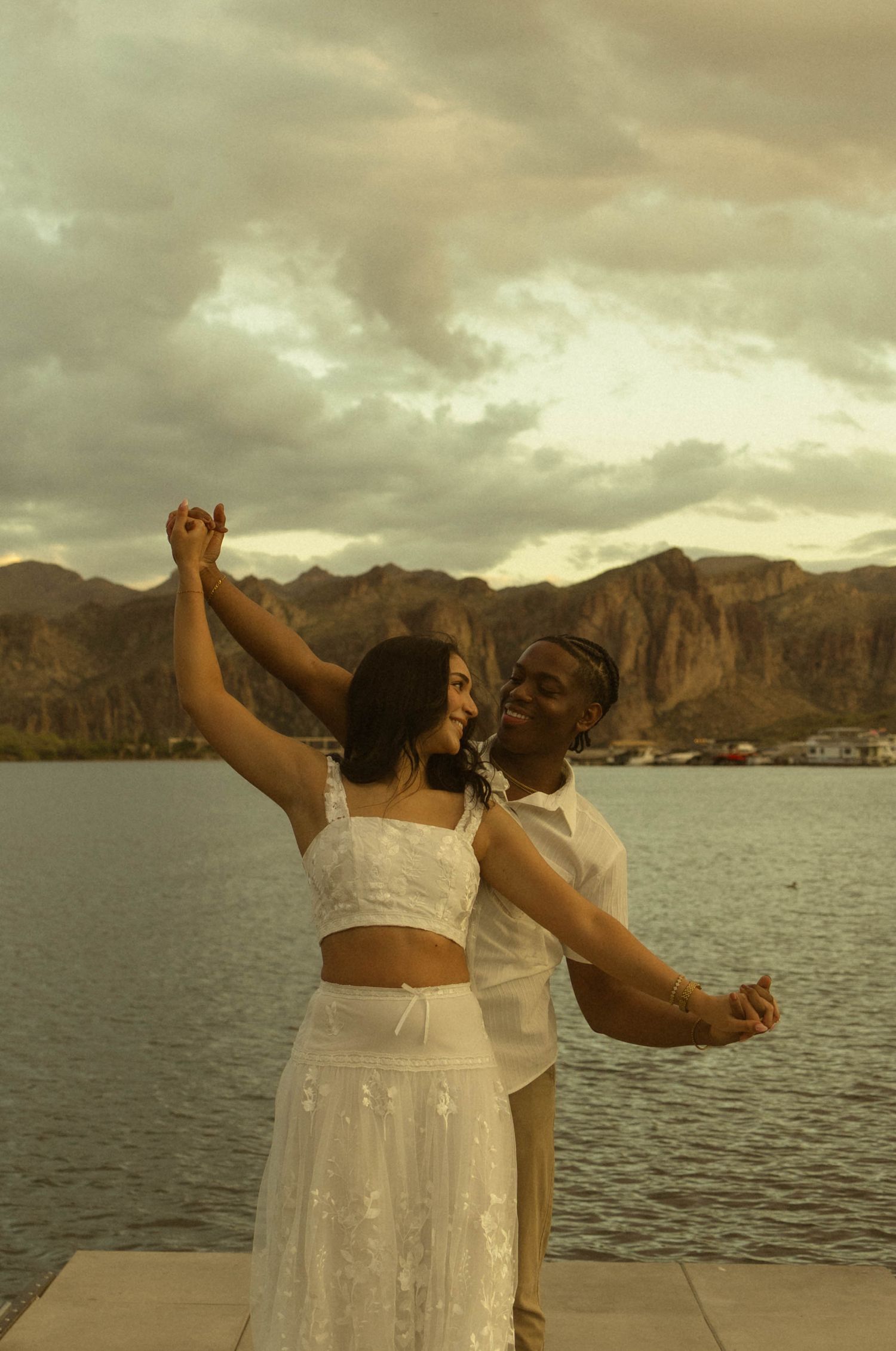 Couple in white outfits dancing by a lake with mountain backdrop at sunset.