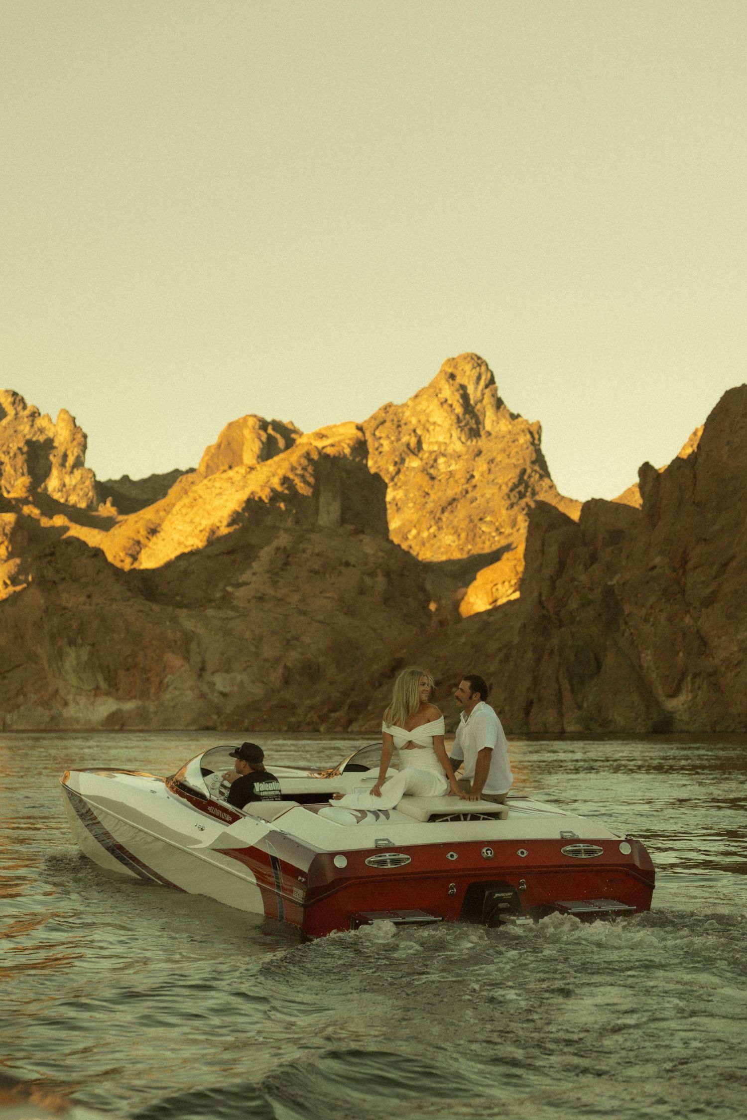 Couple sitting together on a speedboat cruising on calm water with dramatic sunlit mountain cliffs behind.