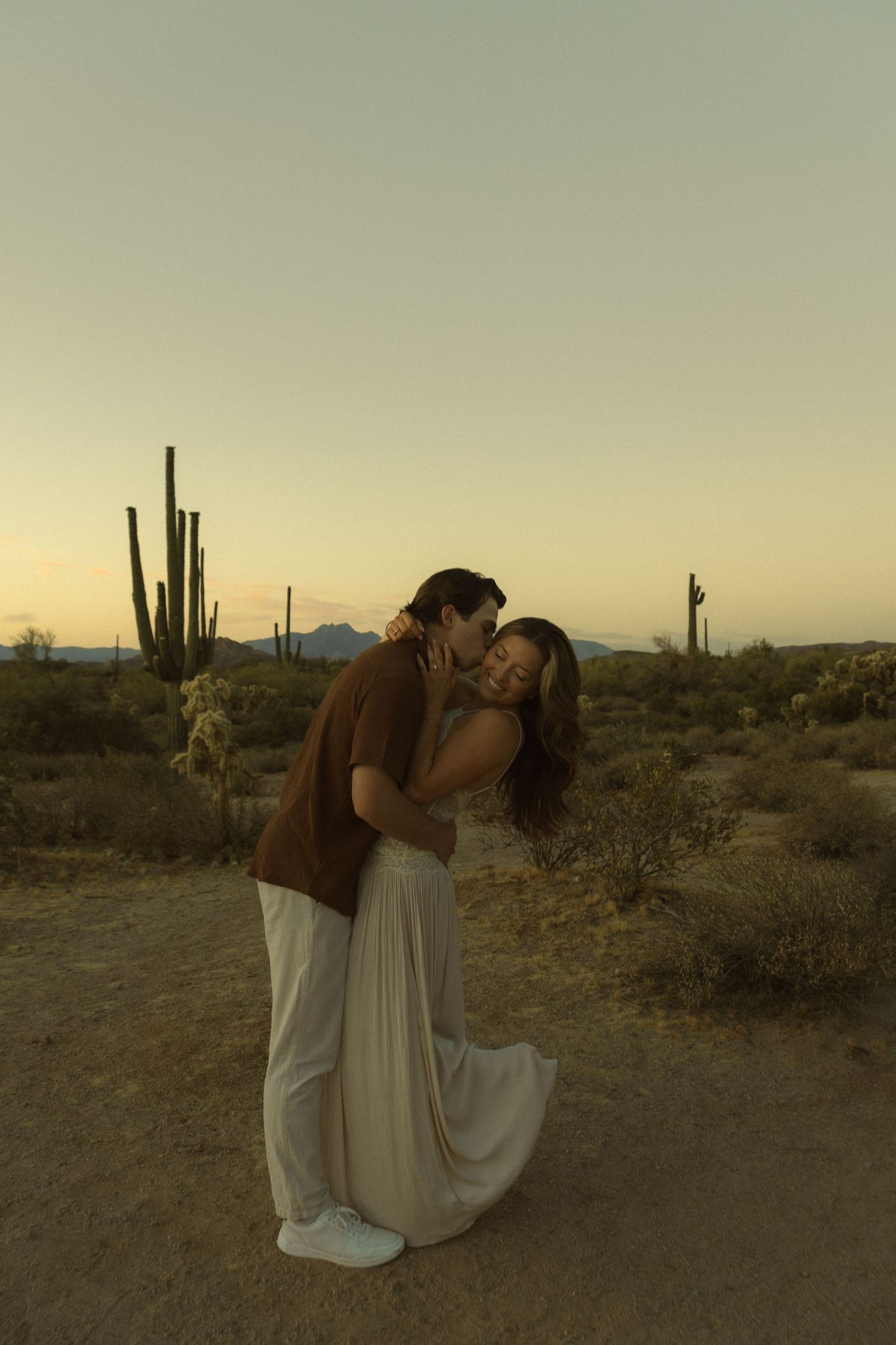 Couple embracing tenderly among saguaro cacti in Arizona desert at sunset.