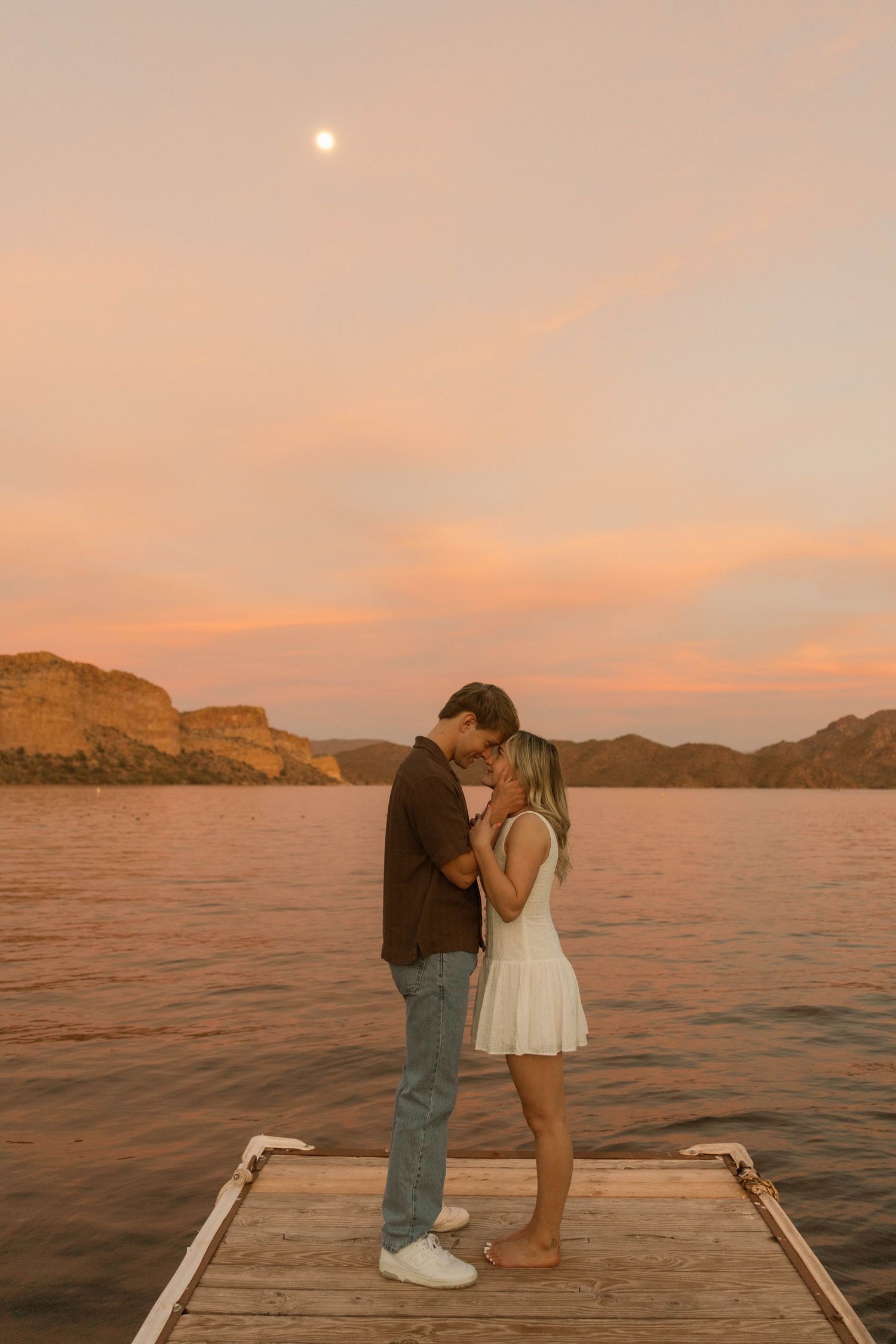 A couple stands together on a wooden dock during sunset with mountains and water in the background.