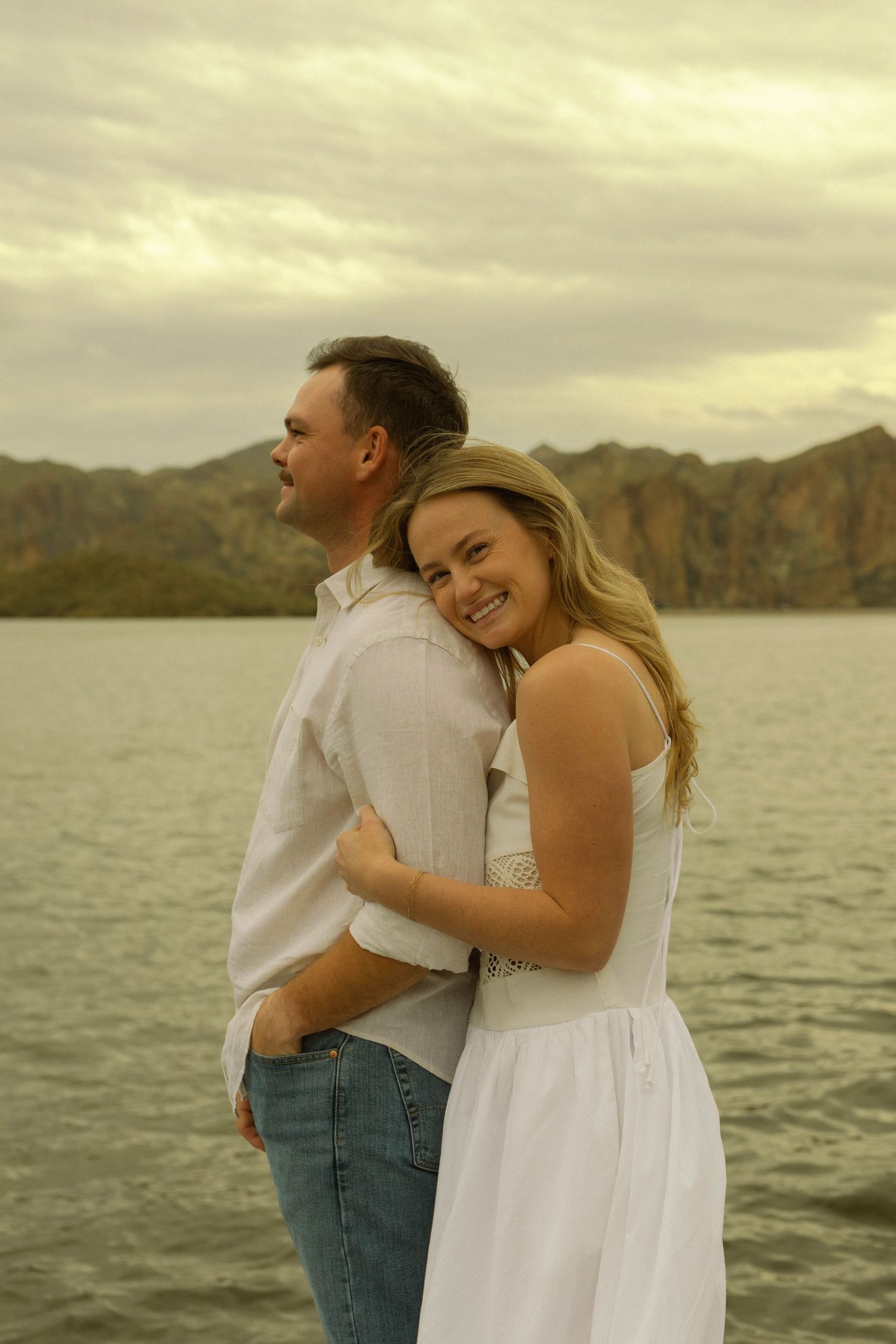 A couple shares a romantic moment on the beach at sunset with mountains and water in the background.