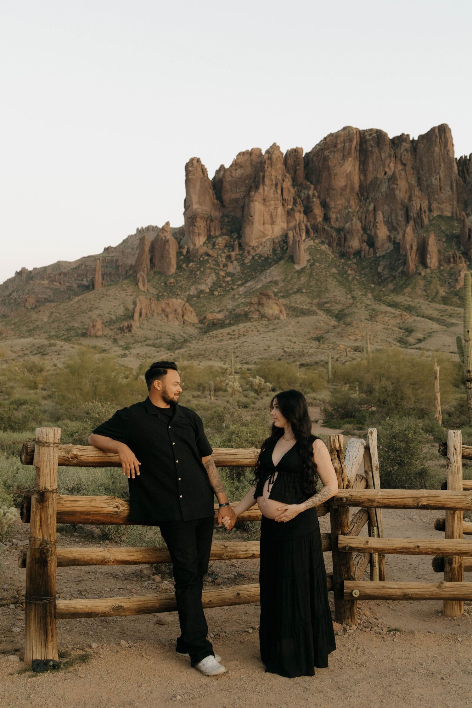 A couple leans against a wooden fence with the Superstition Mountains rising dramatically behind them.