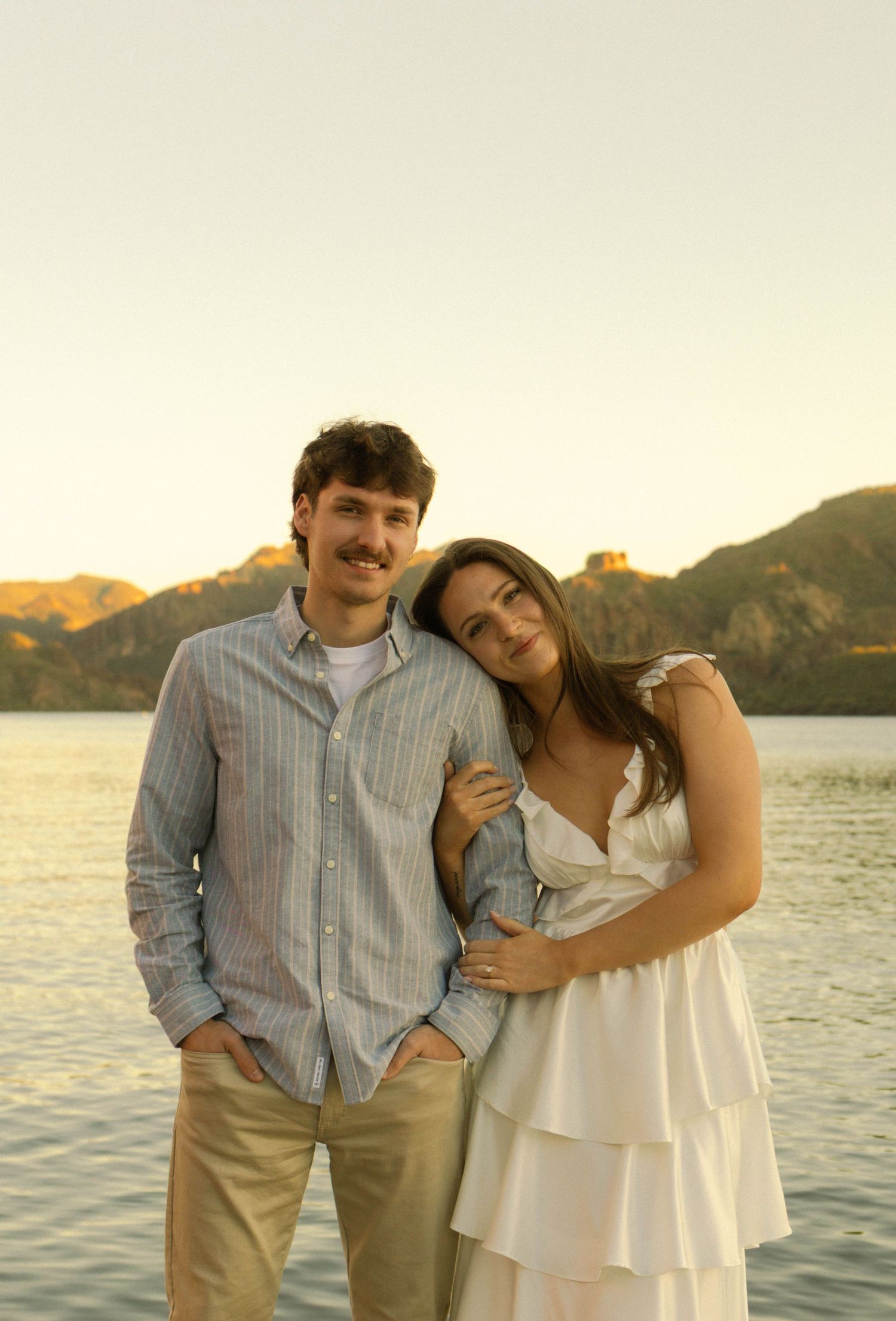 A couple shares a romantic moment at sunset by a lake with mountains in the background.