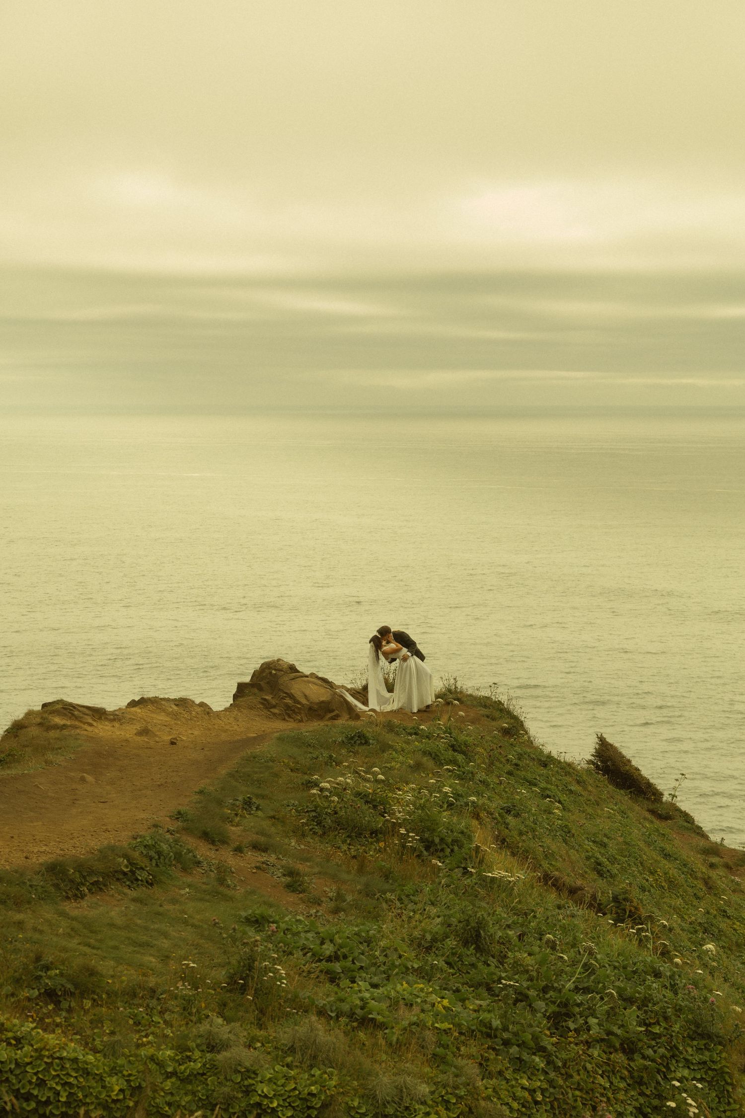 A lone figure in white stands on a rugged coastal cliff overlooking a calm, misty ocean under overcast skies.
