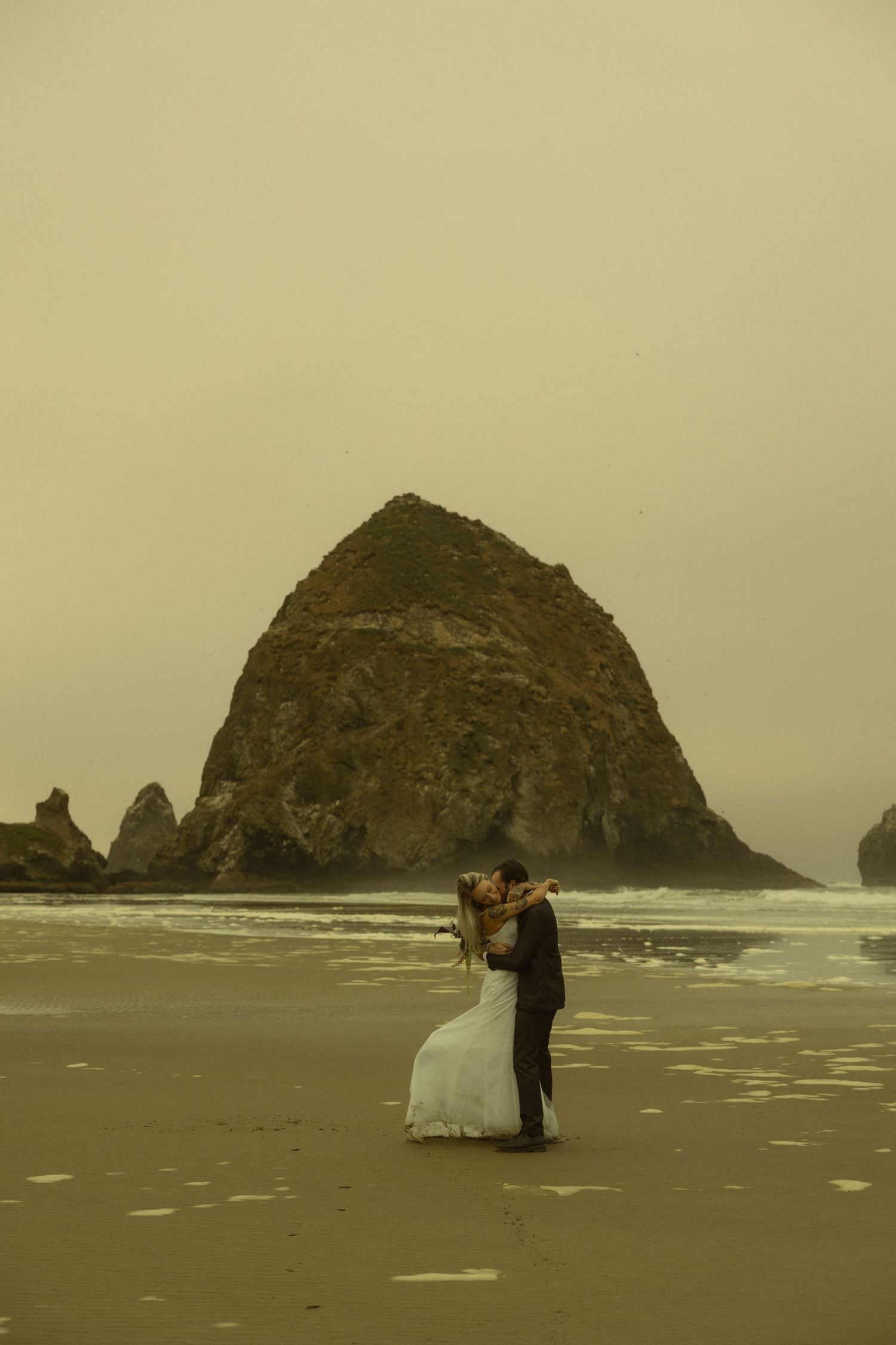 Wedding couple embraces on the sandy shore with iconic Haystack Rock rising from the ocean at Cannon Beach, Oregon.