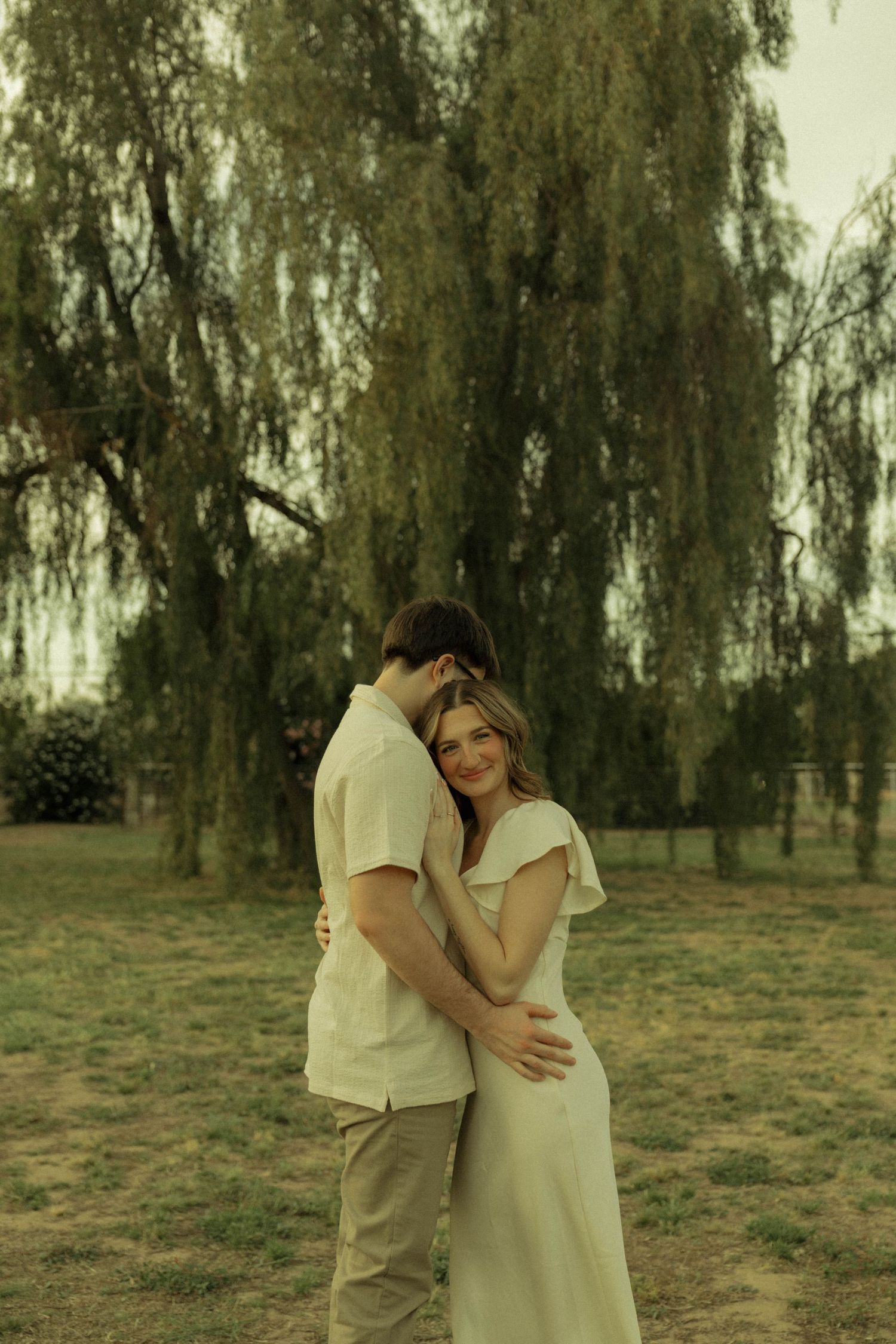 Couple embracing tenderly in front of a weeping willow tree, woman in white dress smiling softly outdoors.