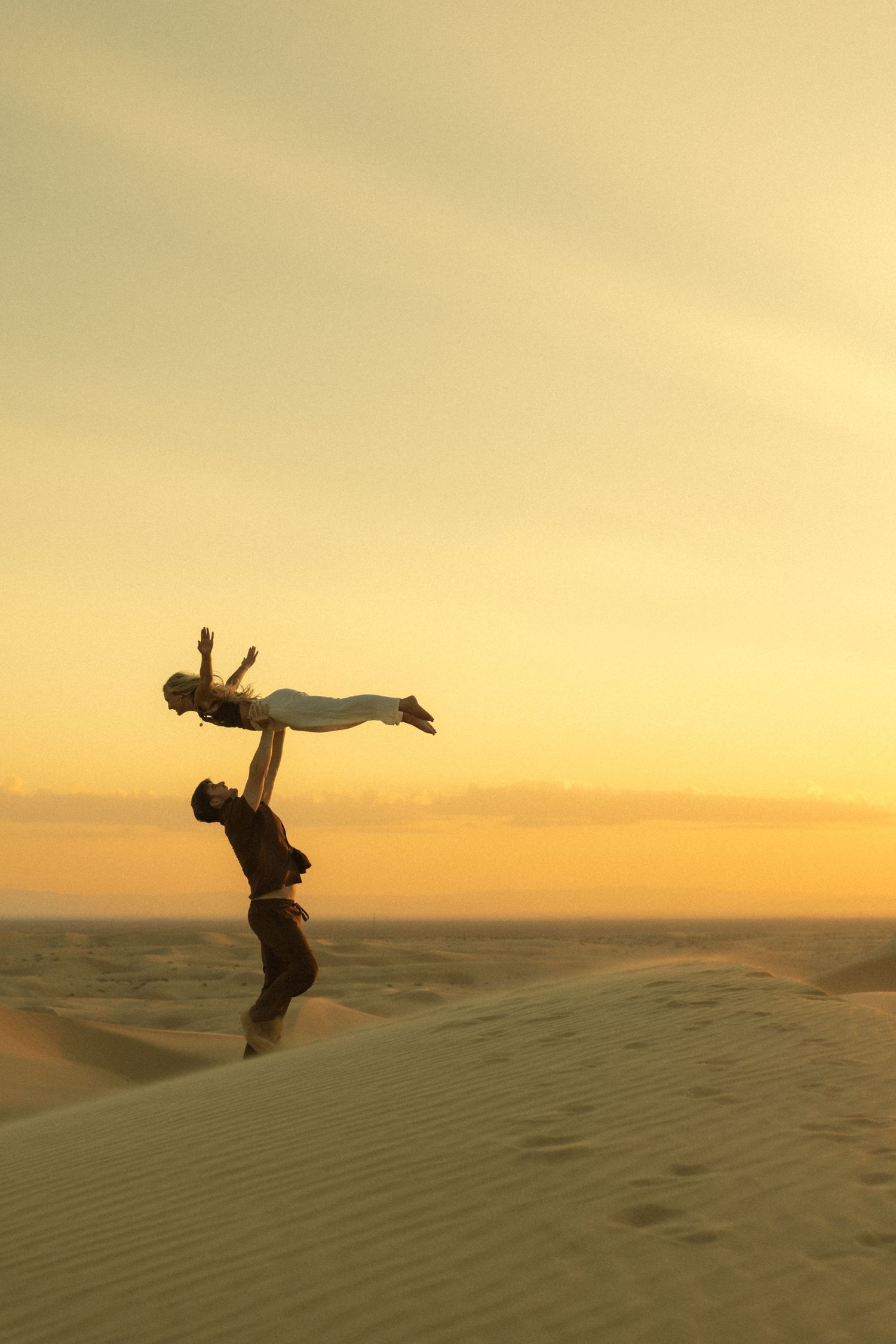Silhouette of man lifting woman overhead on desert sand dunes against a dramatic golden sunset sky.