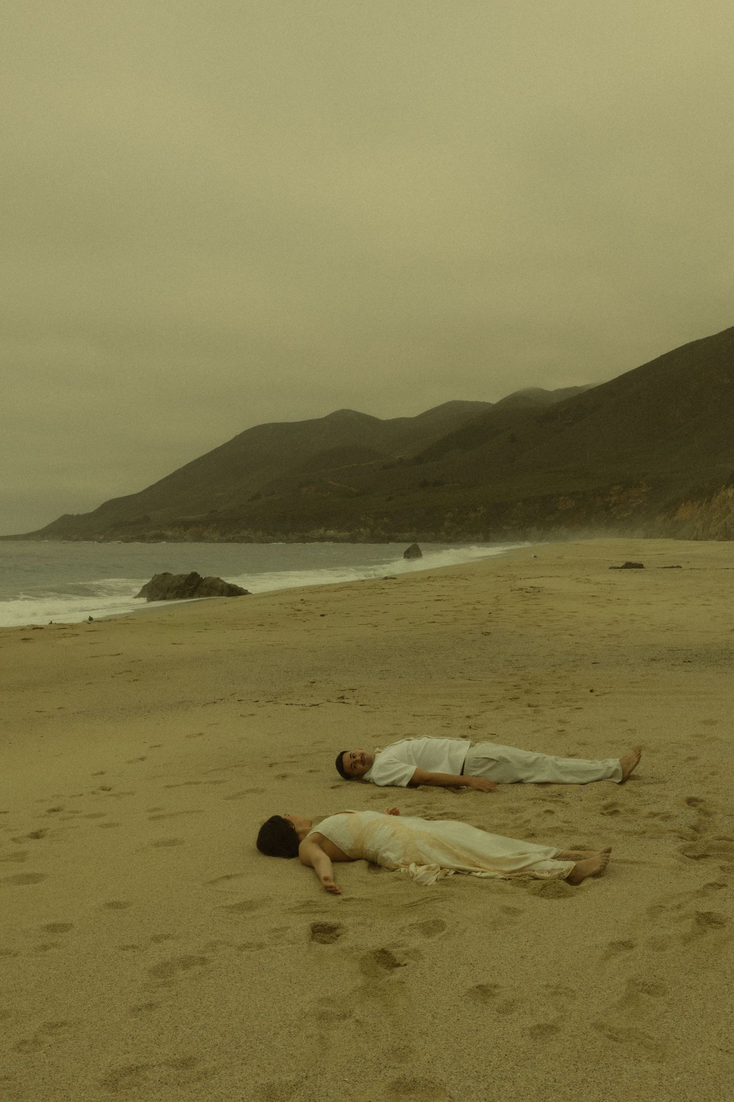 Two people in white clothing lying on a sandy beach with mountains and ocean in the background.