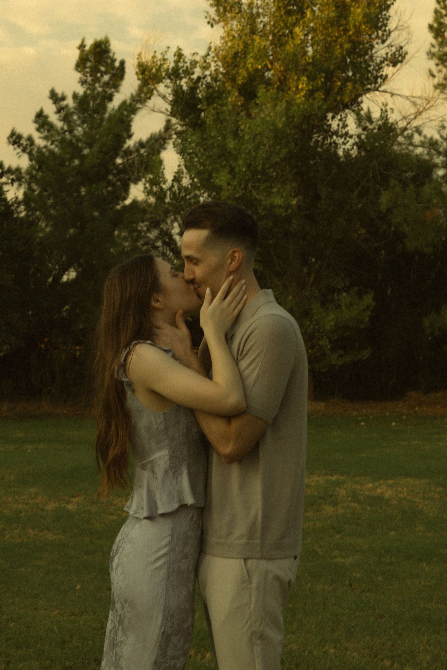 A couple shares a romantic kiss outdoors on a green lawn with lush trees in the background at golden hour.