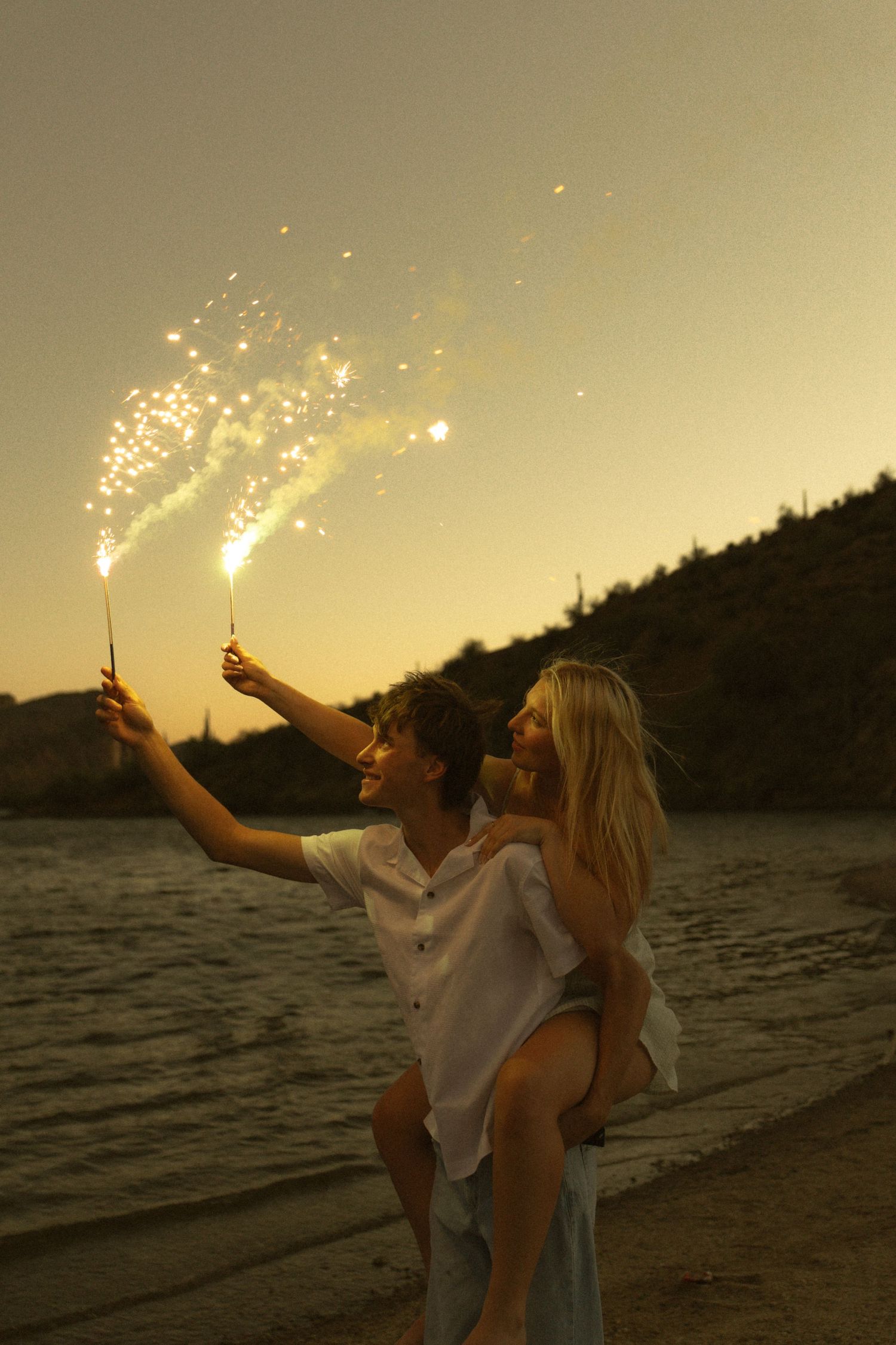 Couple holding sparklers on a beach at sunset, man giving woman a piggyback ride near the water.