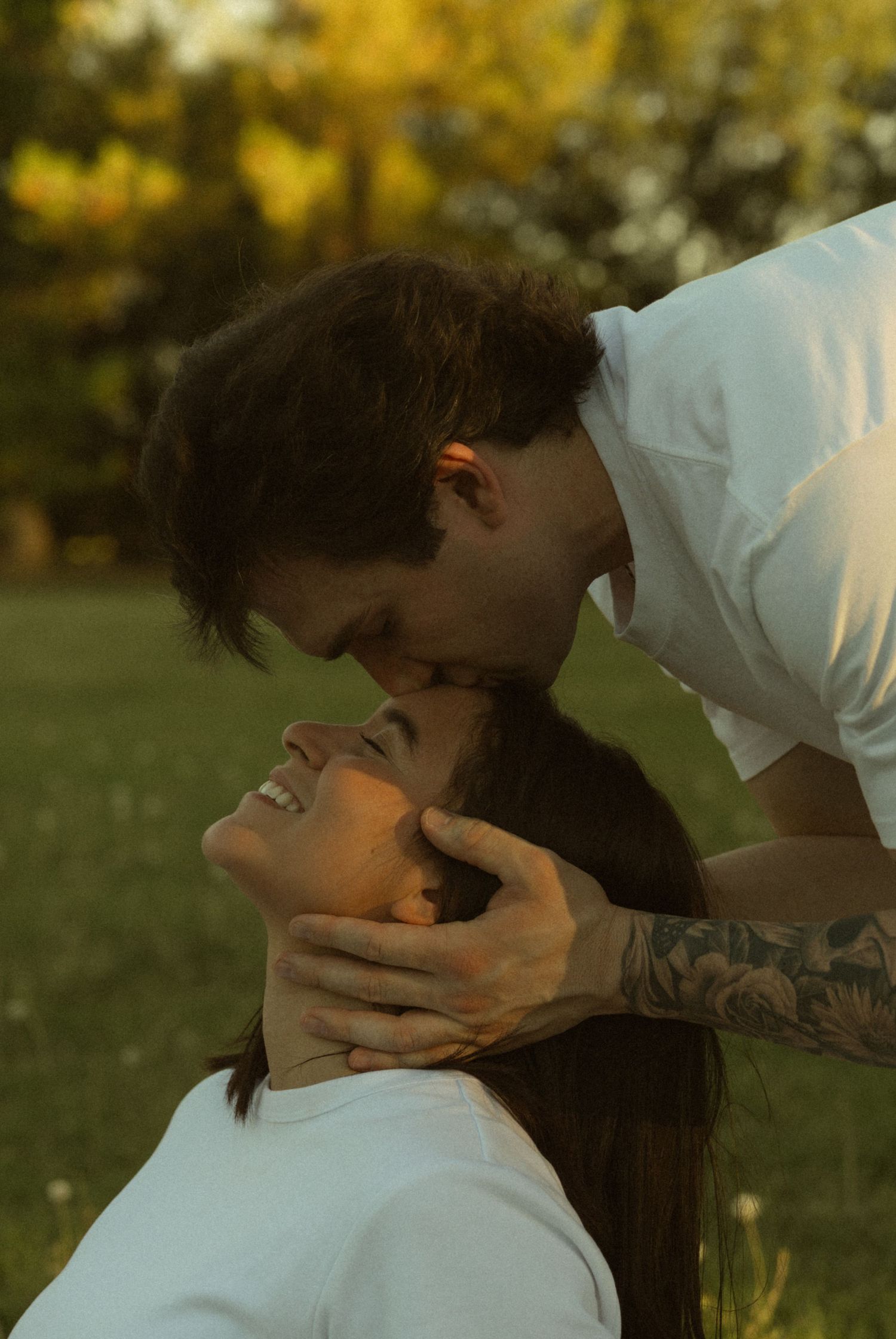 A tattooed man in a white shirt tenderly kisses a smiling woman on the forehead in a lush green park.