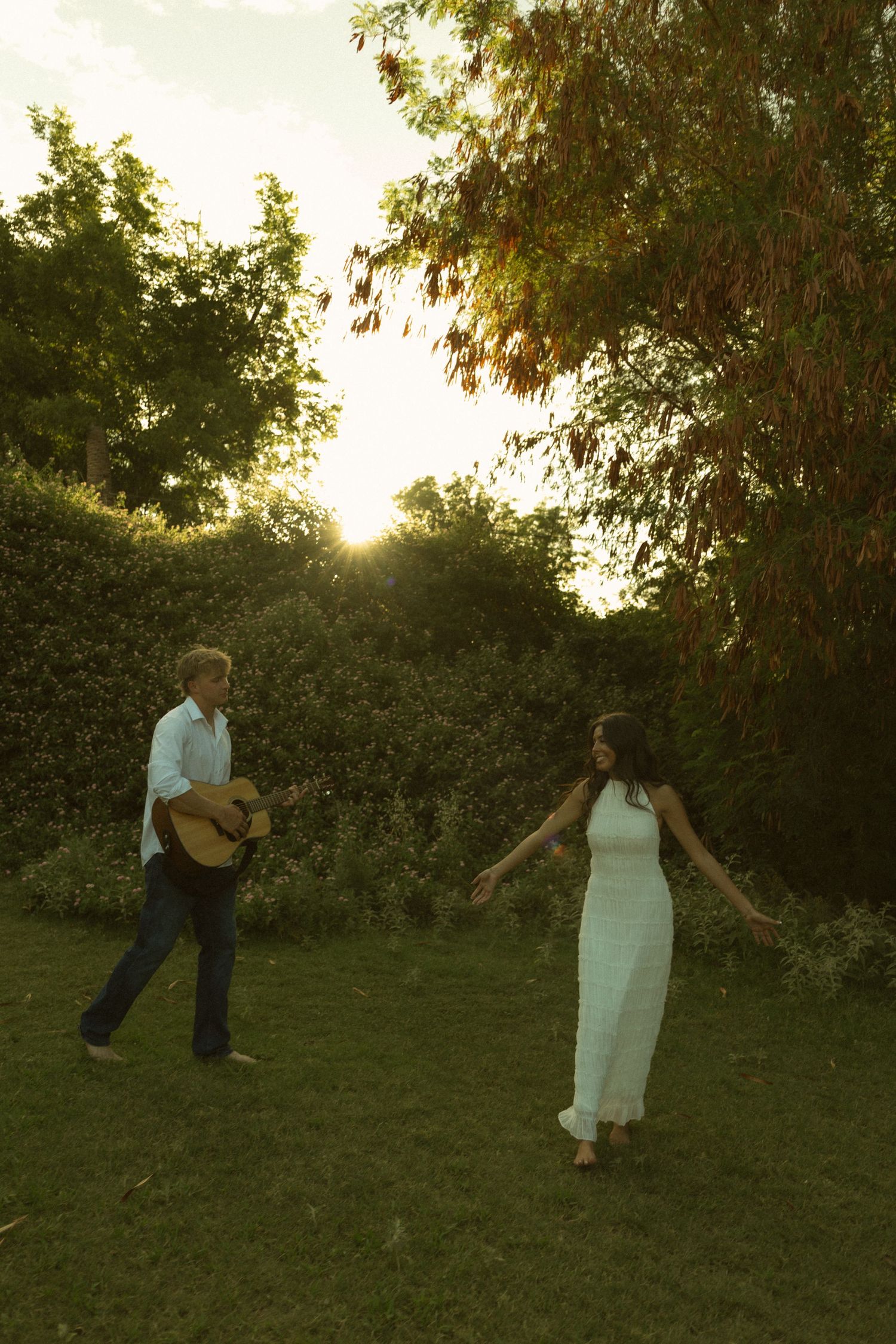Couple dancing in a sunlit garden, man playing acoustic guitar while woman in white dress twirls barefoot on green grass.