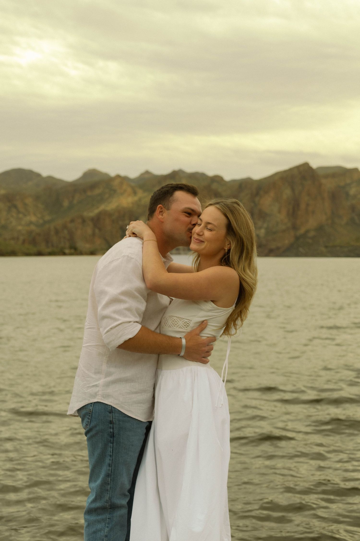 A romantic moment captured at sunset by a lake with rugged mountains in the background.