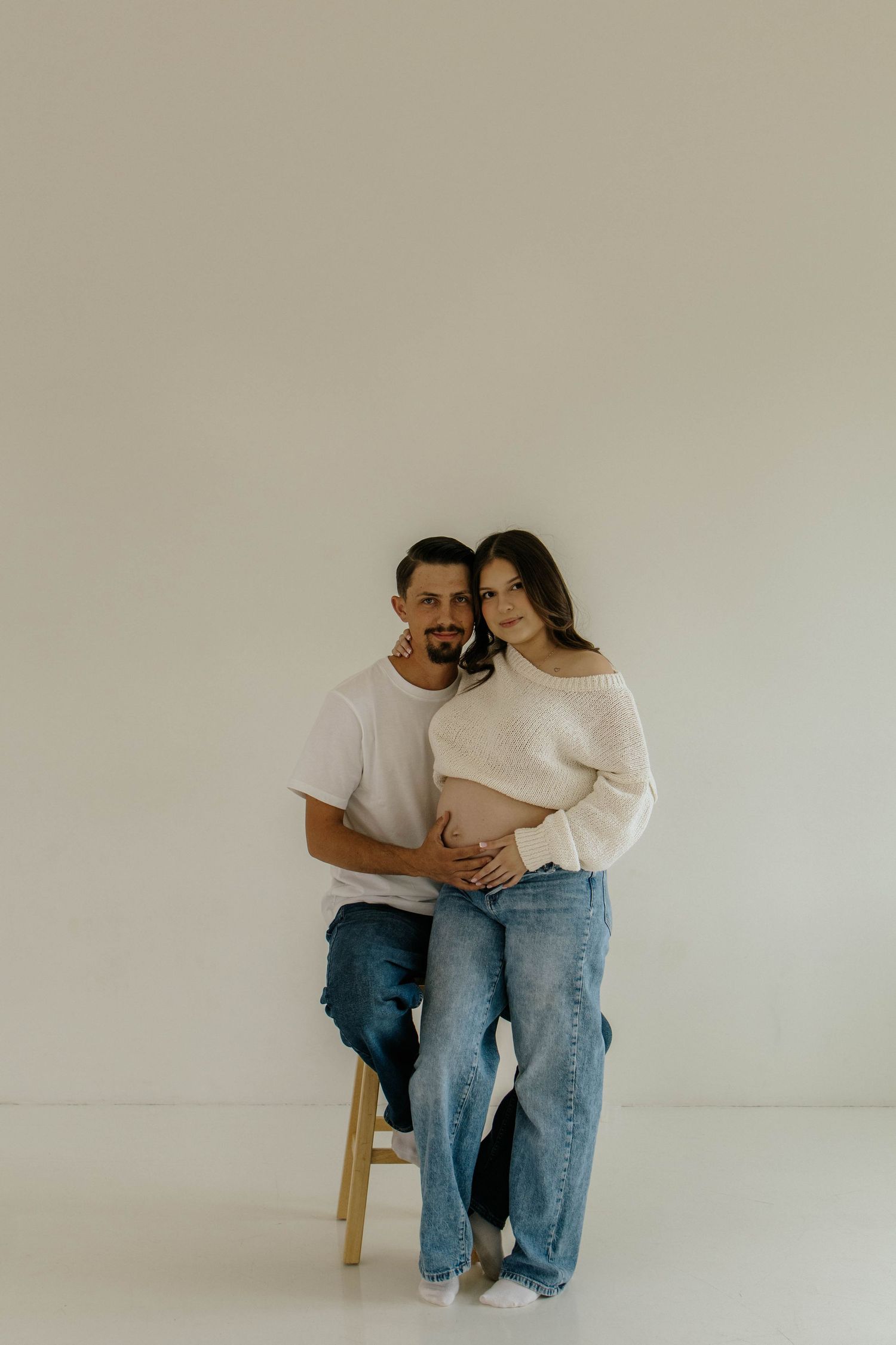 A minimalist maternity photoshoot with a couple sitting on a wooden stool against a beige wall.