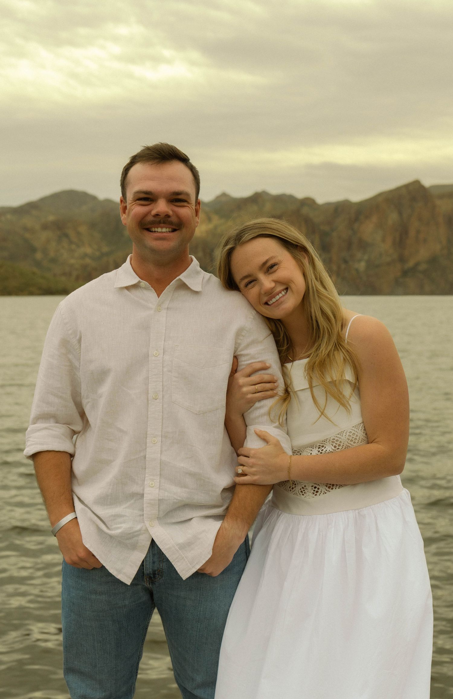 A happy couple in white outfits poses together against a scenic mountain lake backdrop.