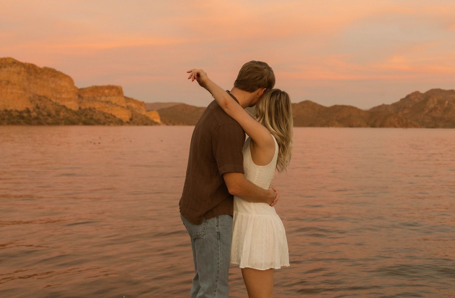 A couple embraces at sunset by a serene lake surrounded by mountains in warm golden light.