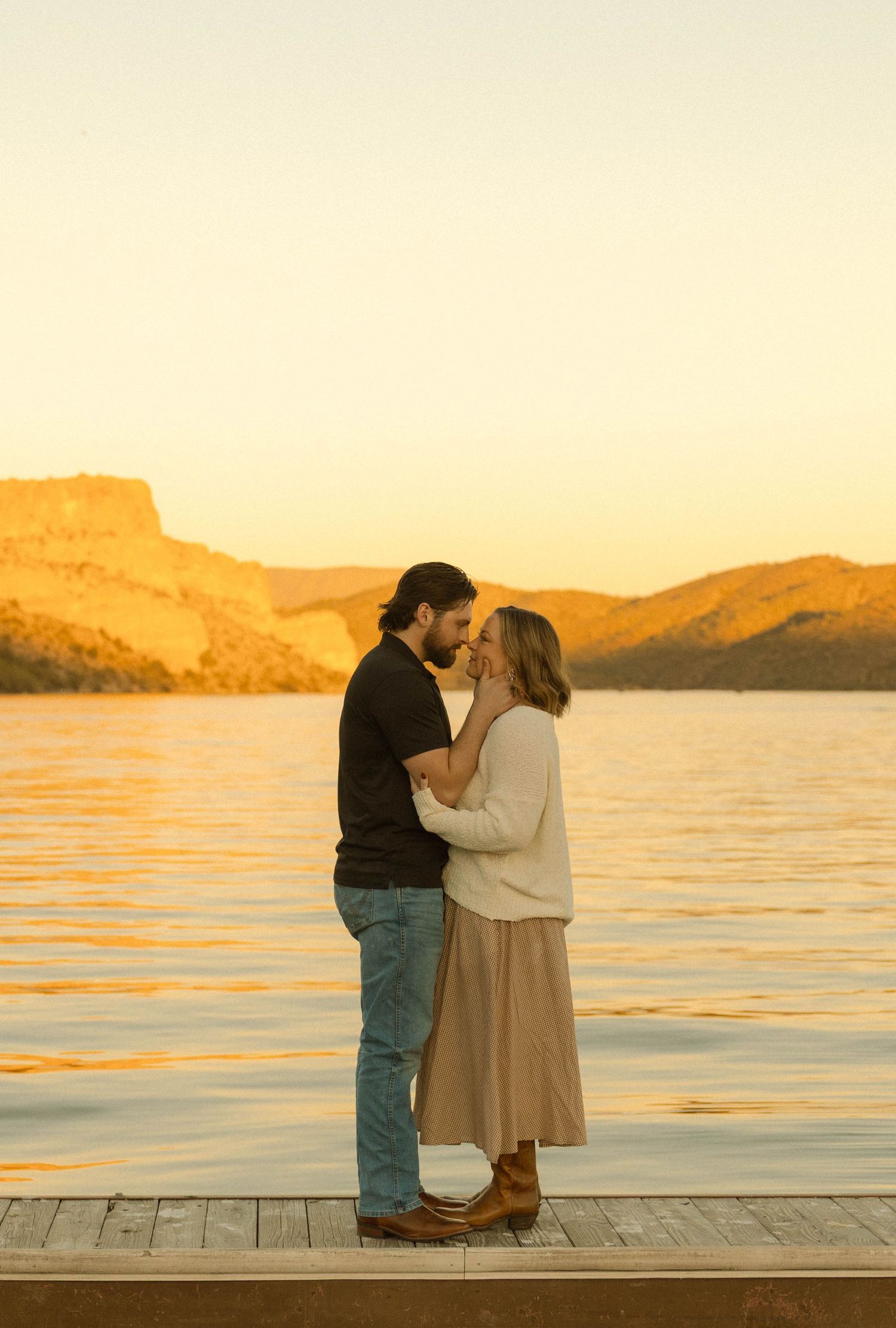 A couple shares a romantic moment on a wooden dock during golden hour sunset over a serene lake with mountains in background.